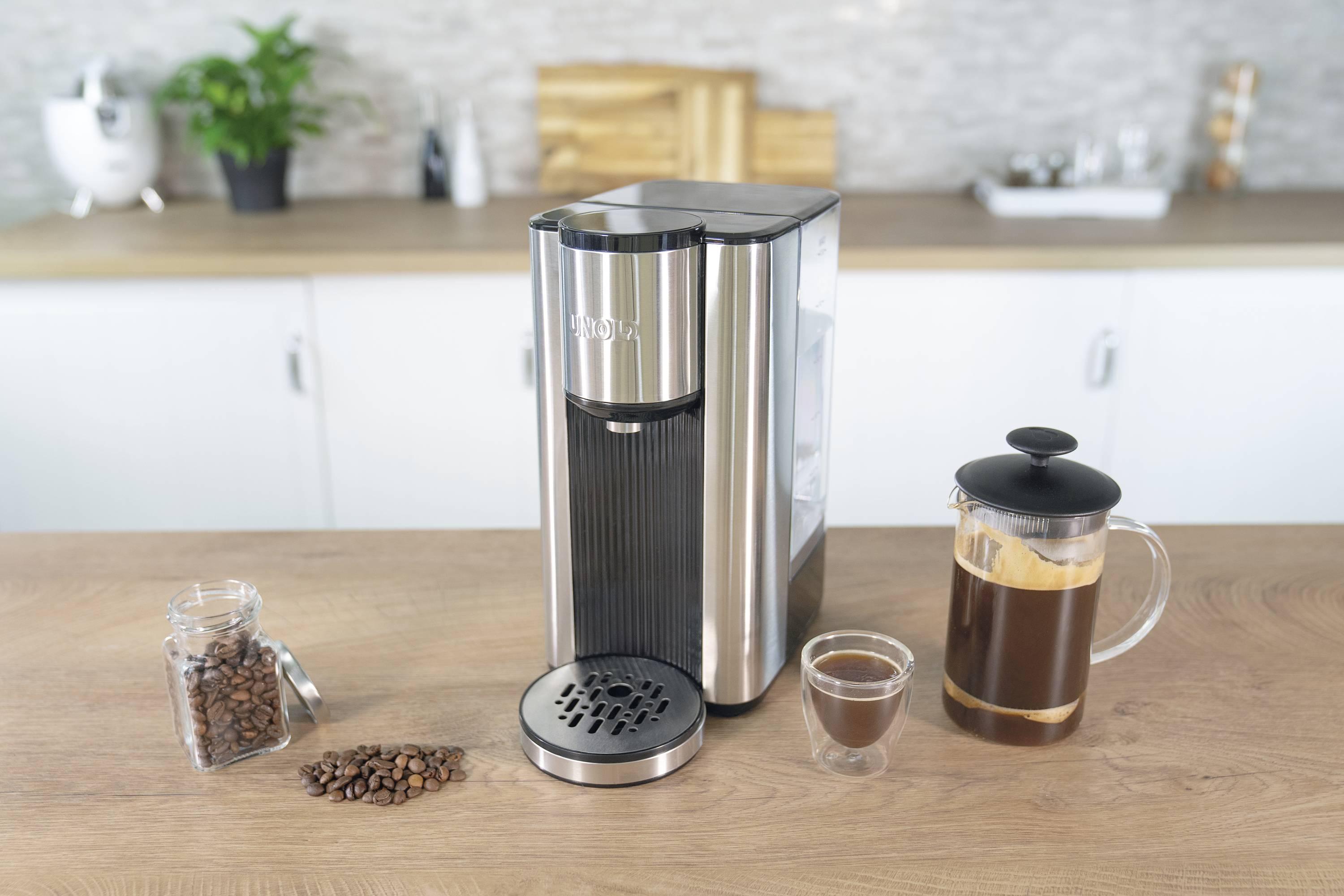 Eine silberne Kaffeemaschine steht auf einer Küchenarbeitsplatte, umgeben von einem Glas Kaffee, Kaffeebohnen und einer Kaffeepresse.