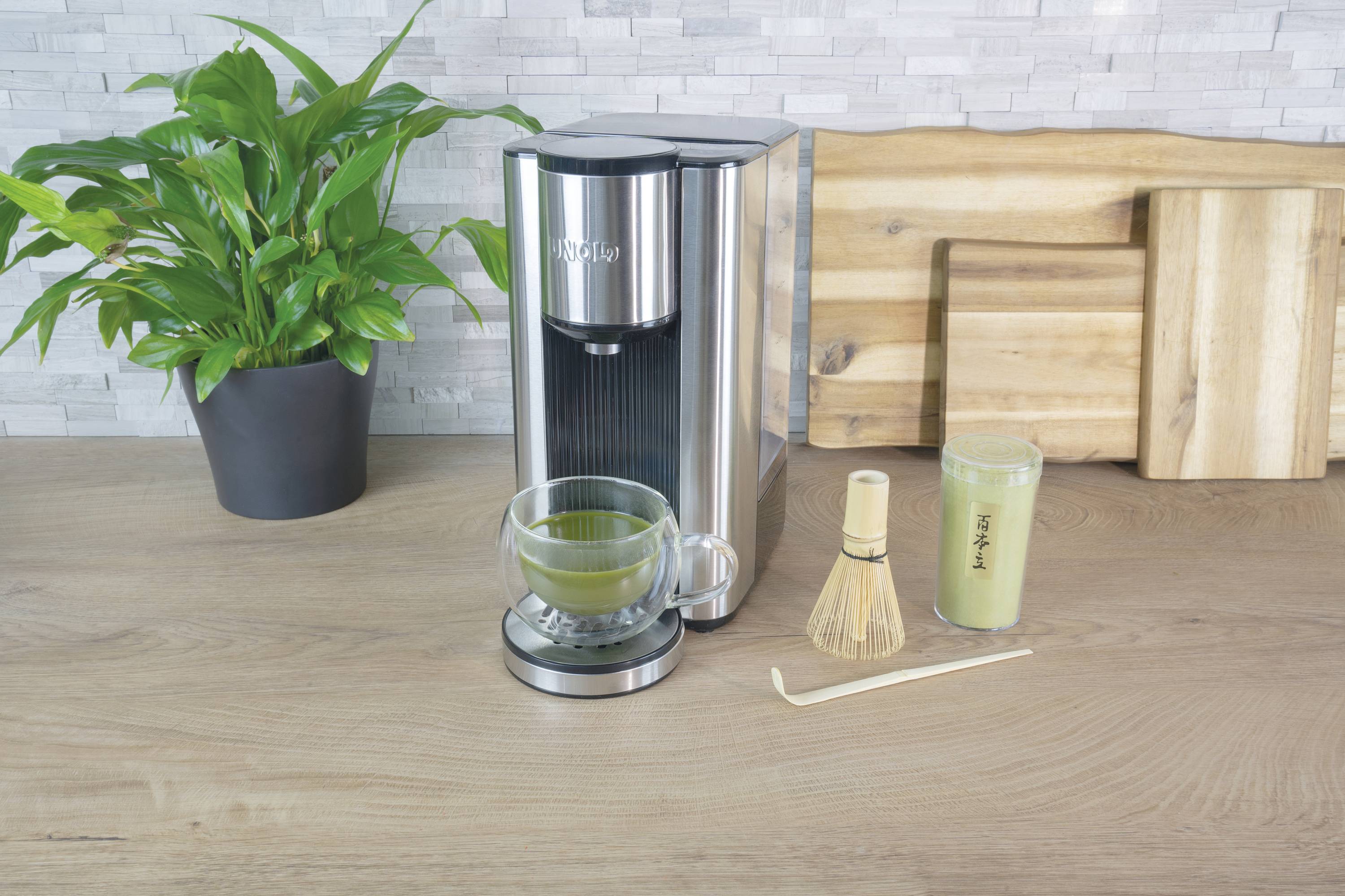 Matcha-Maschine mit Glas, Matcha-Pulver, Bambusbesen und Kanne auf Holztisch. Pflanze und Schneidebretter im Hintergrund.
