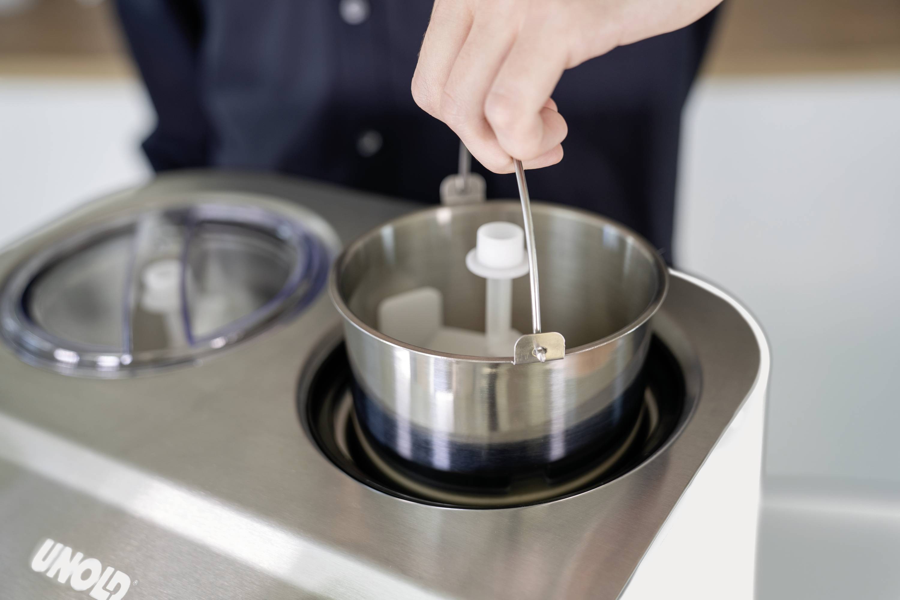 Hand hält den Rührstab eines Eismakers über die geöffnete Edelstahlschüssel der Maschine.