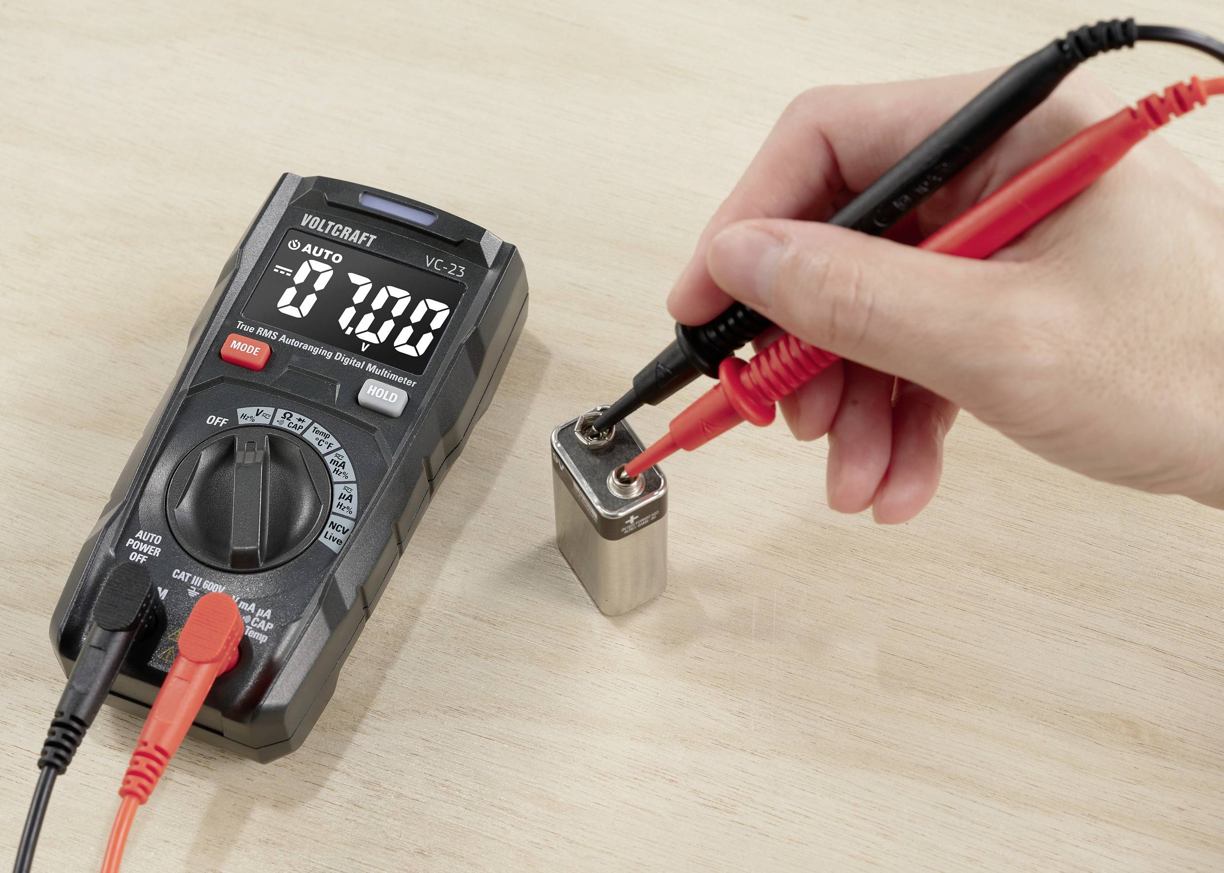 Hand mit Prüfspitzen eines Multimeters misst Spannung einer 9-Volt-Batterie. Multimeter zeigt 7,00 Volt. Holzoberfläche im Hintergrund.