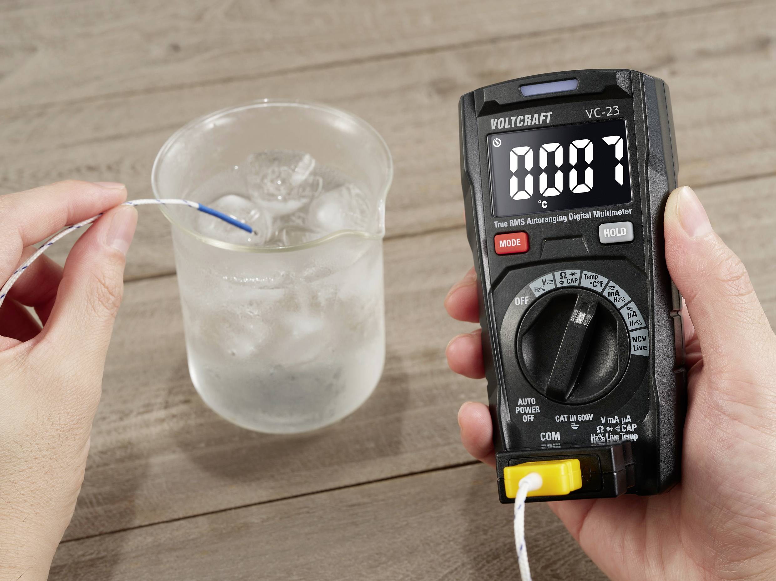 Eine Person misst die Temperatur des Wassers in einem Becherglas mit einem Digitalmultimeter, das 0,07 °C anzeigt.