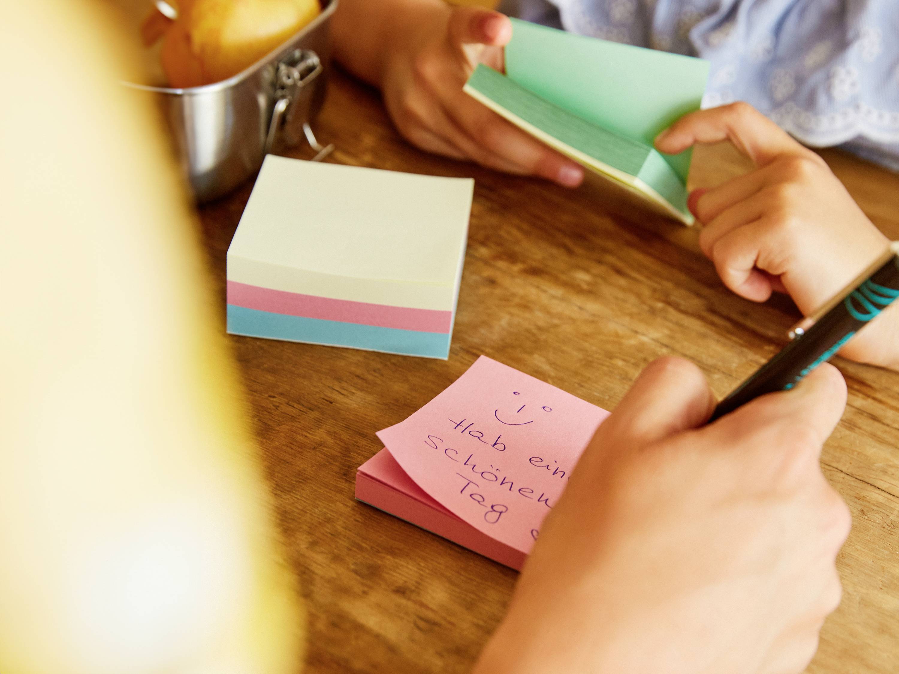 Person schreibt auf einen rosa Notizzettel: 'Hab einen schönen Tag :)'. Weitere farbige Haftnotizen und Hände sind sichtbar.