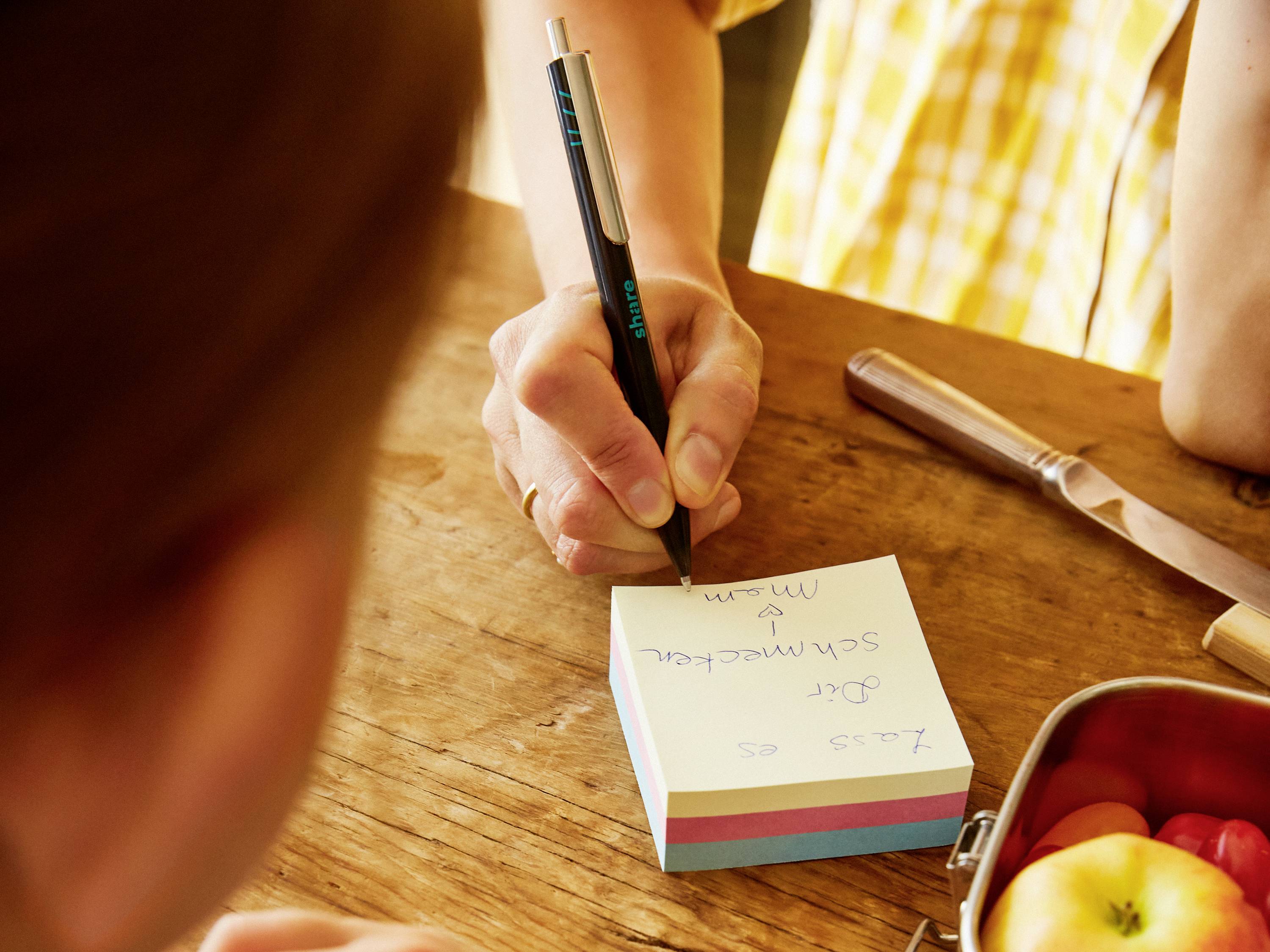 Eine Person schreibt auf einen bunten Notizblock. Auf dem Tisch stehen ein Messer und eine Brotdose mit Obst.