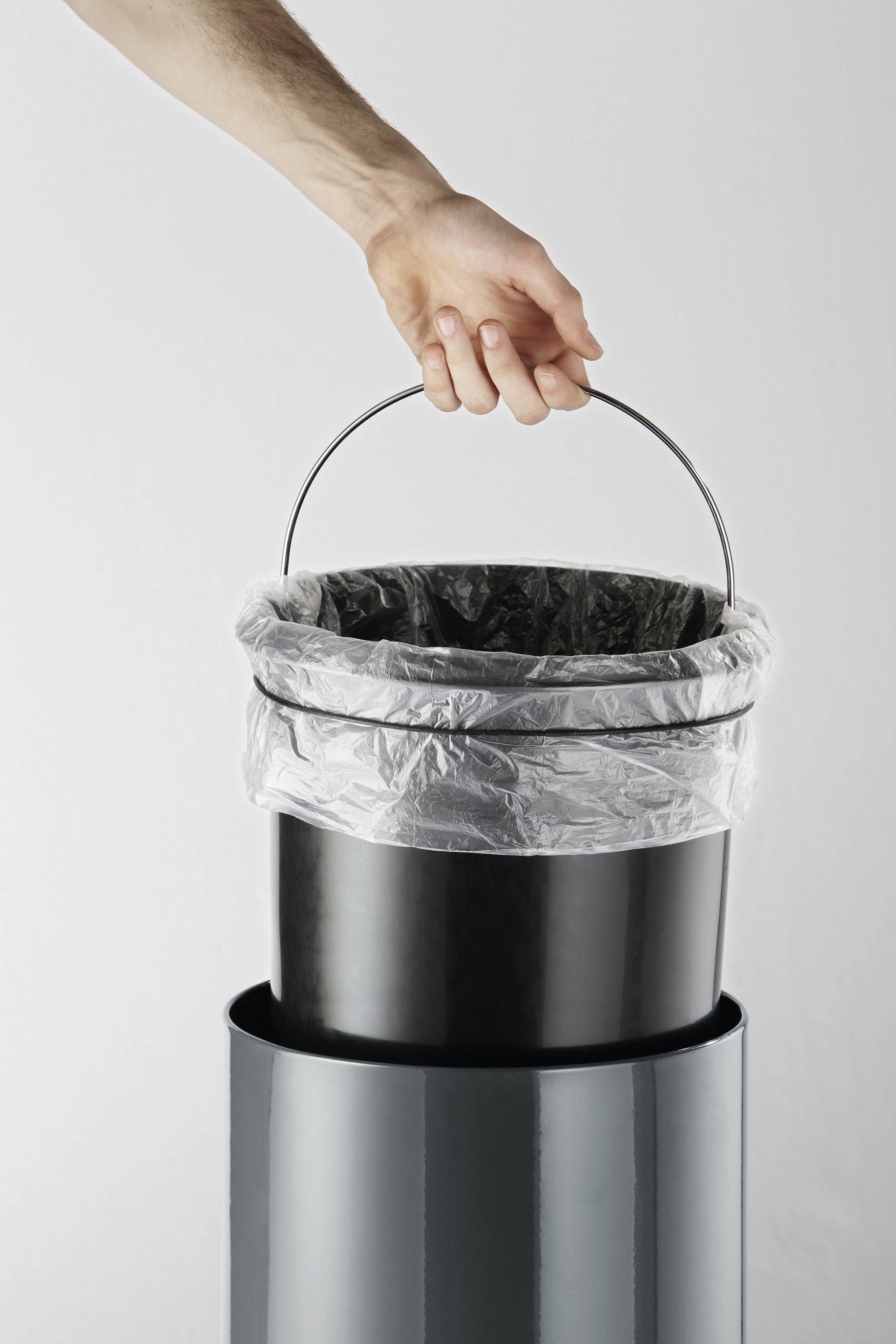 Eine Hand hält einen schwarzen Abfalleimer mit Müllbeutel über einem Metallbehälter.