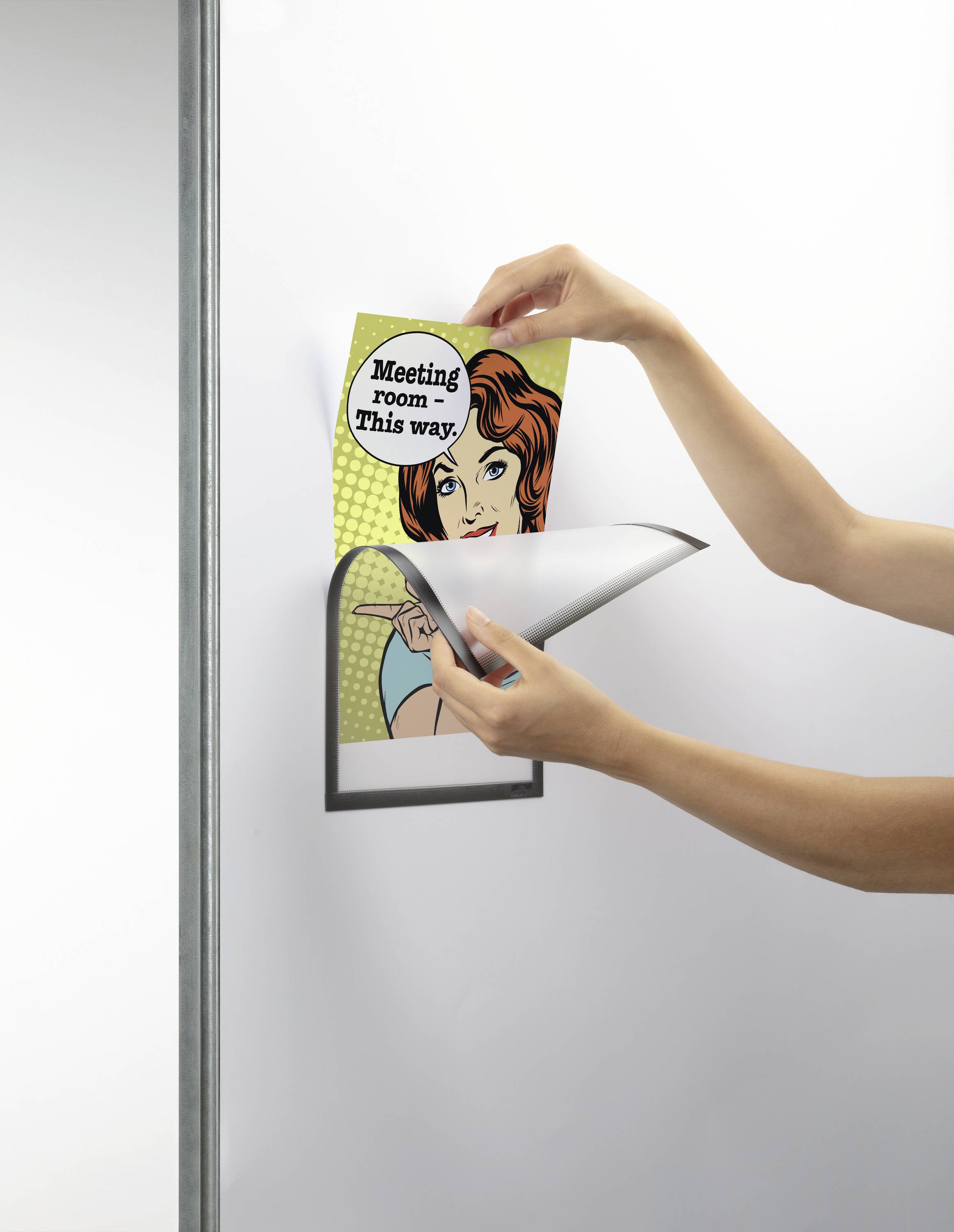Eine Person wechselt ein Poster mit der Aufschrift 'Meeting room this way' in einem Wandrahmen. Das Poster zeigt eine Comicfigur.