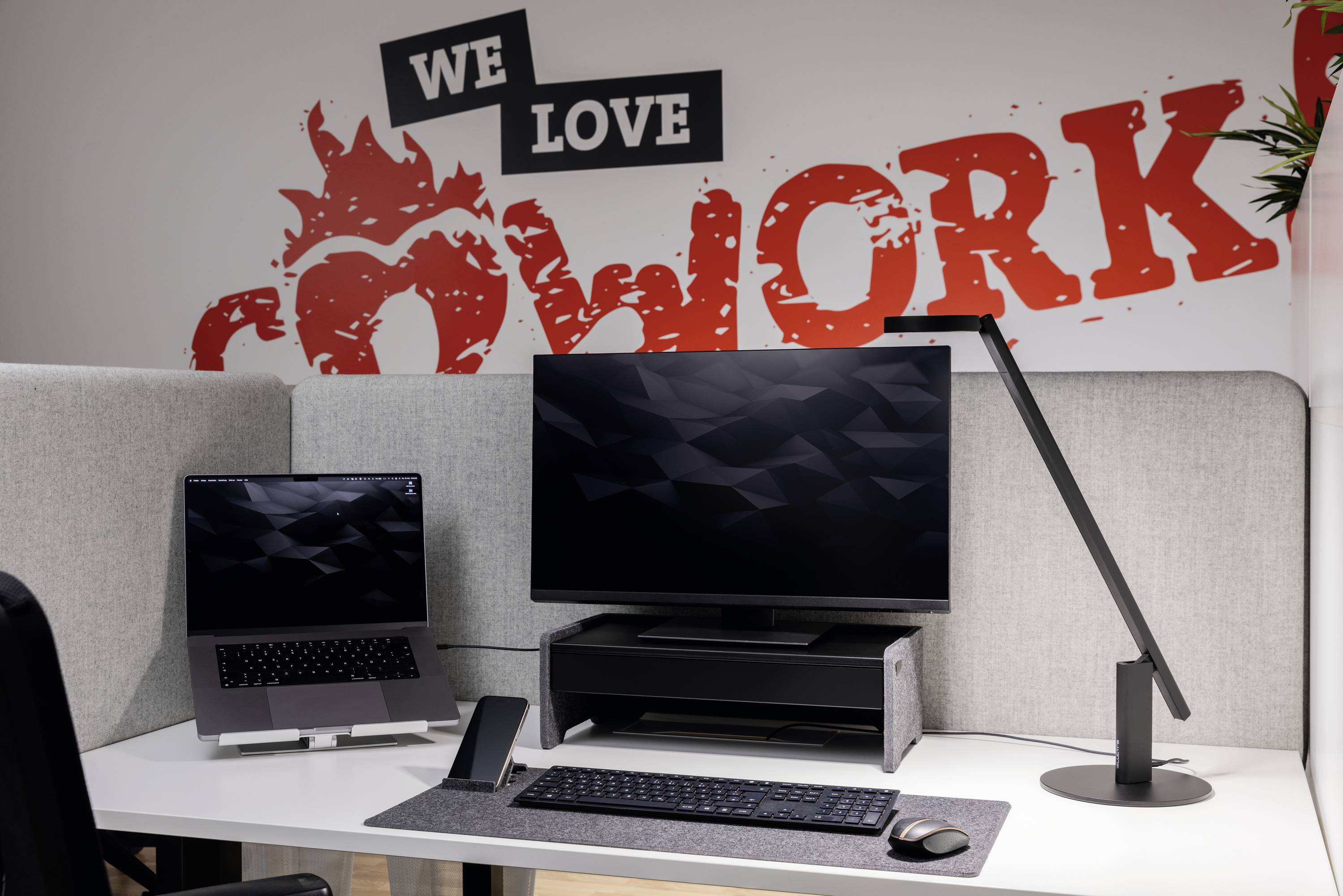 Ein moderner Arbeitsplatz mit einem Laptop, Monitor, Lampe und Aufschrift 'We love cowork' an der Wand im Hintergrund.