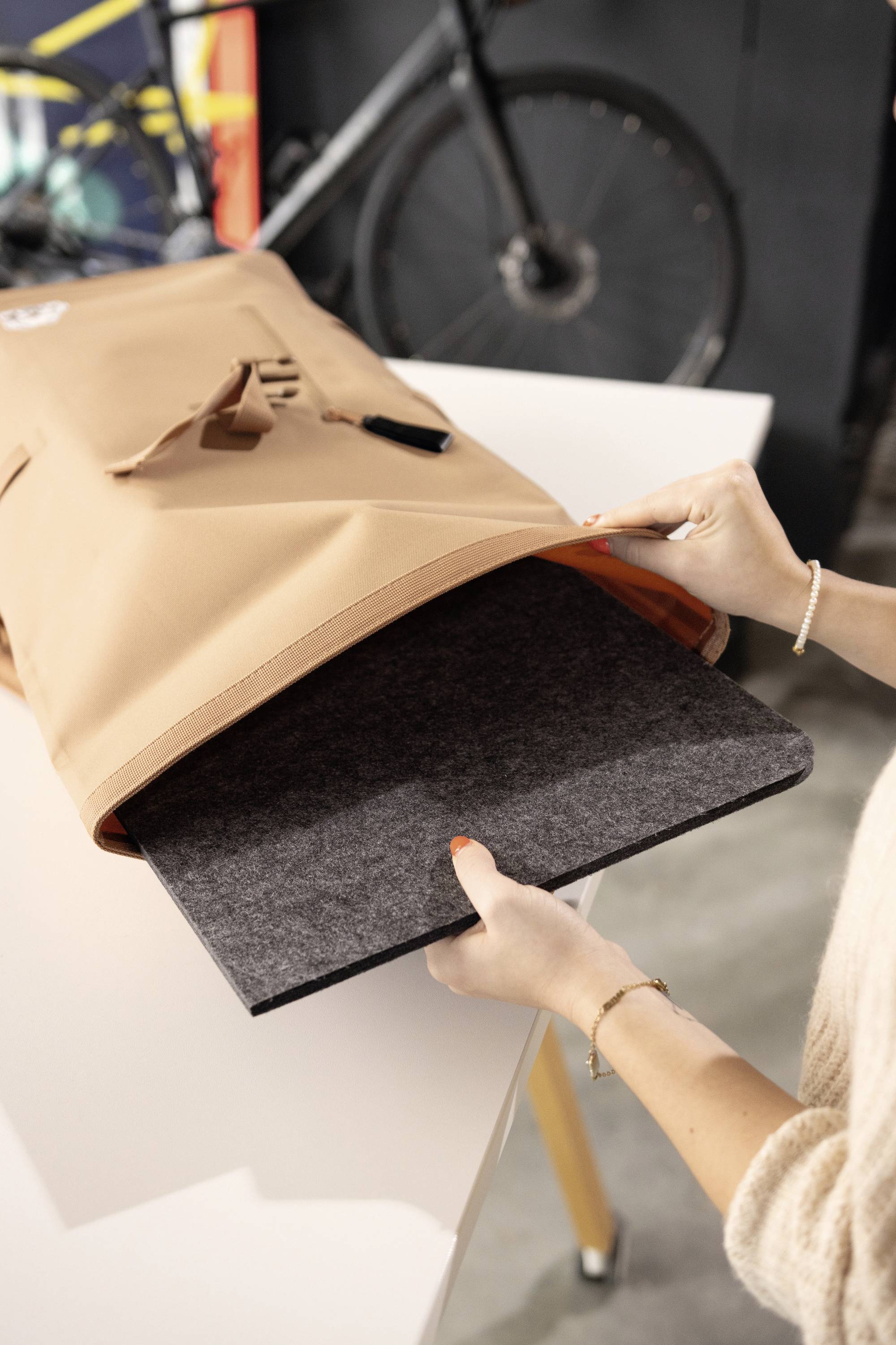 Person packt einen Laptop in eine beige Tasche auf einem Tisch. Ein Fahrrad ist im Hintergrund sichtbar.