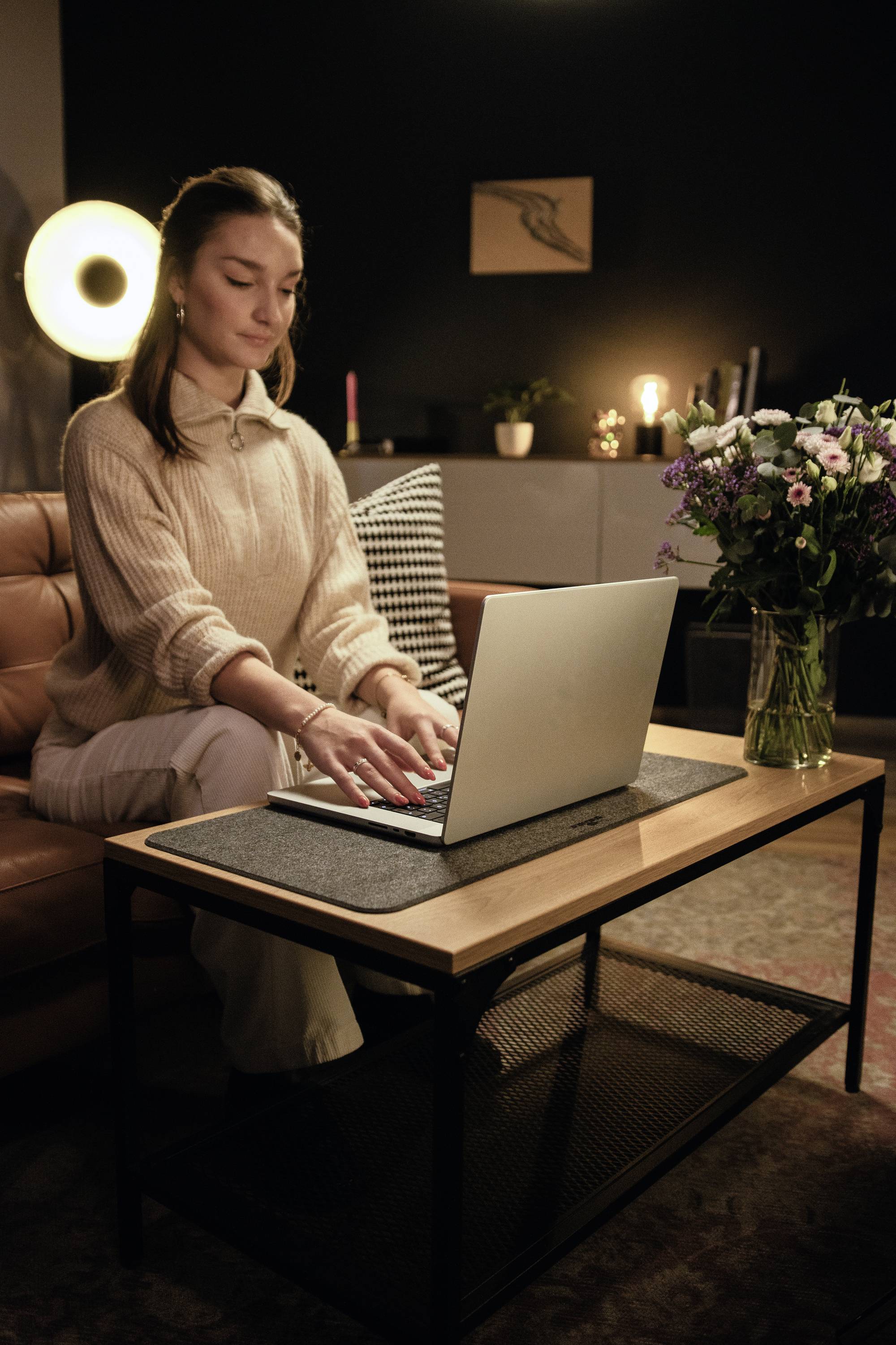 Frau sitzt auf einem Sofa und tippt auf einem Laptop; Blumenstrauß auf dem Tisch daneben. Wohnzimmer mit gedimmtem Licht im Hintergrund.