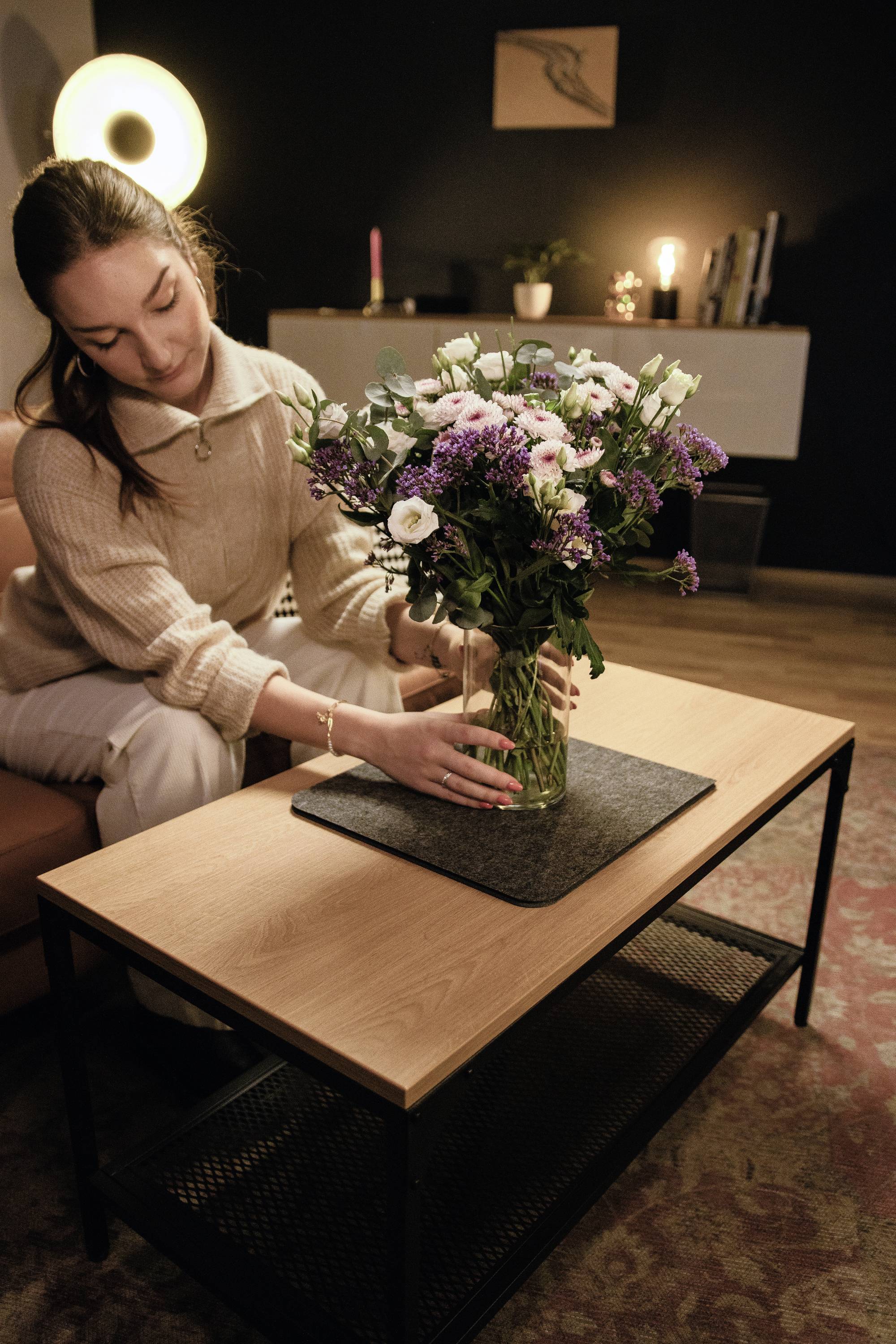 Eine Frau stellt einen Blumenstrauß auf einen Wohnzimmertisch. Im Hintergrund sind eine Lampe und Dekorationen zu sehen.