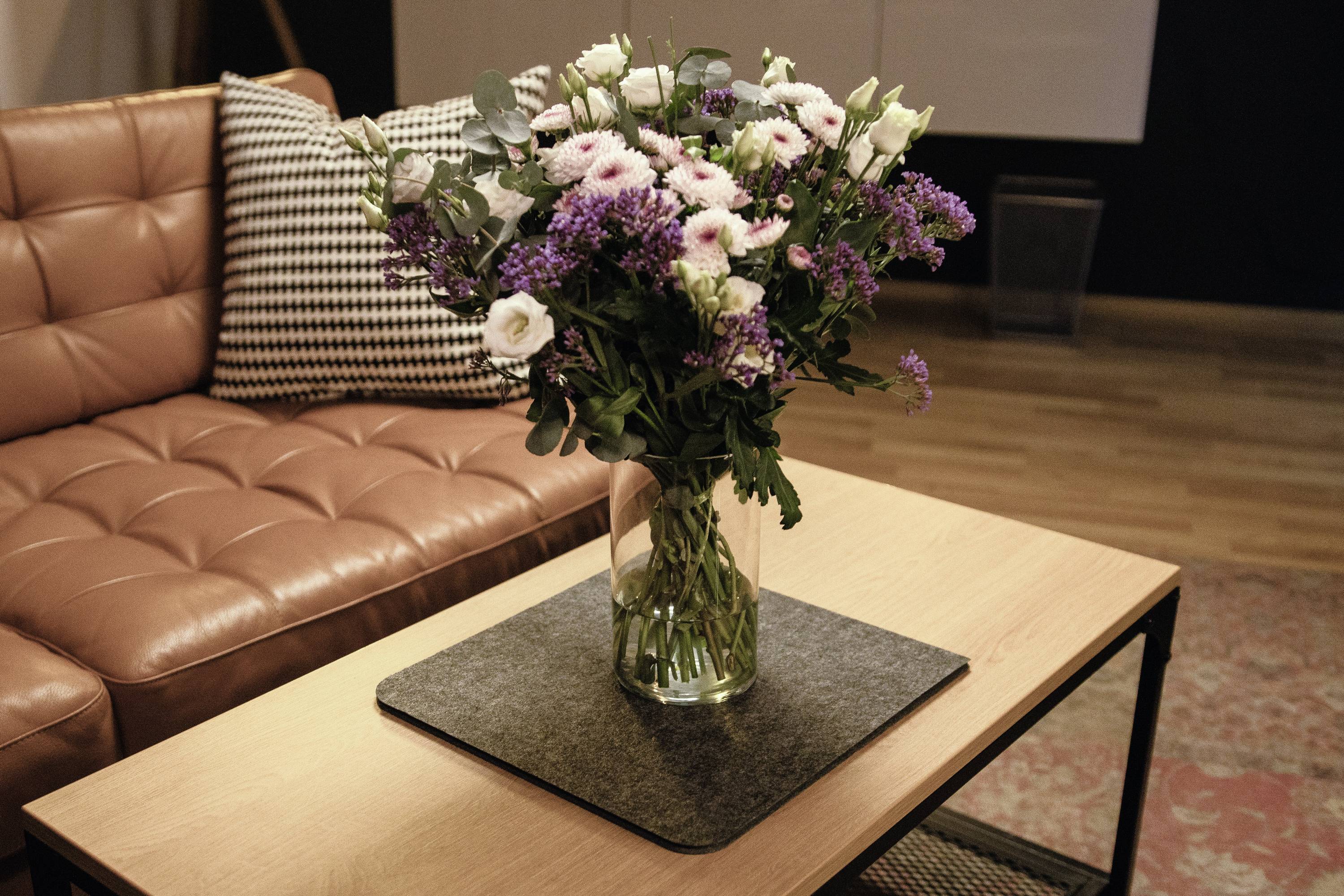 Blumenstrauß mit lila und weißen Blüten in einer Glasvase auf einem Kaffee-Tisch vor einem braunen Ledersofa, im Wohnzimmer.