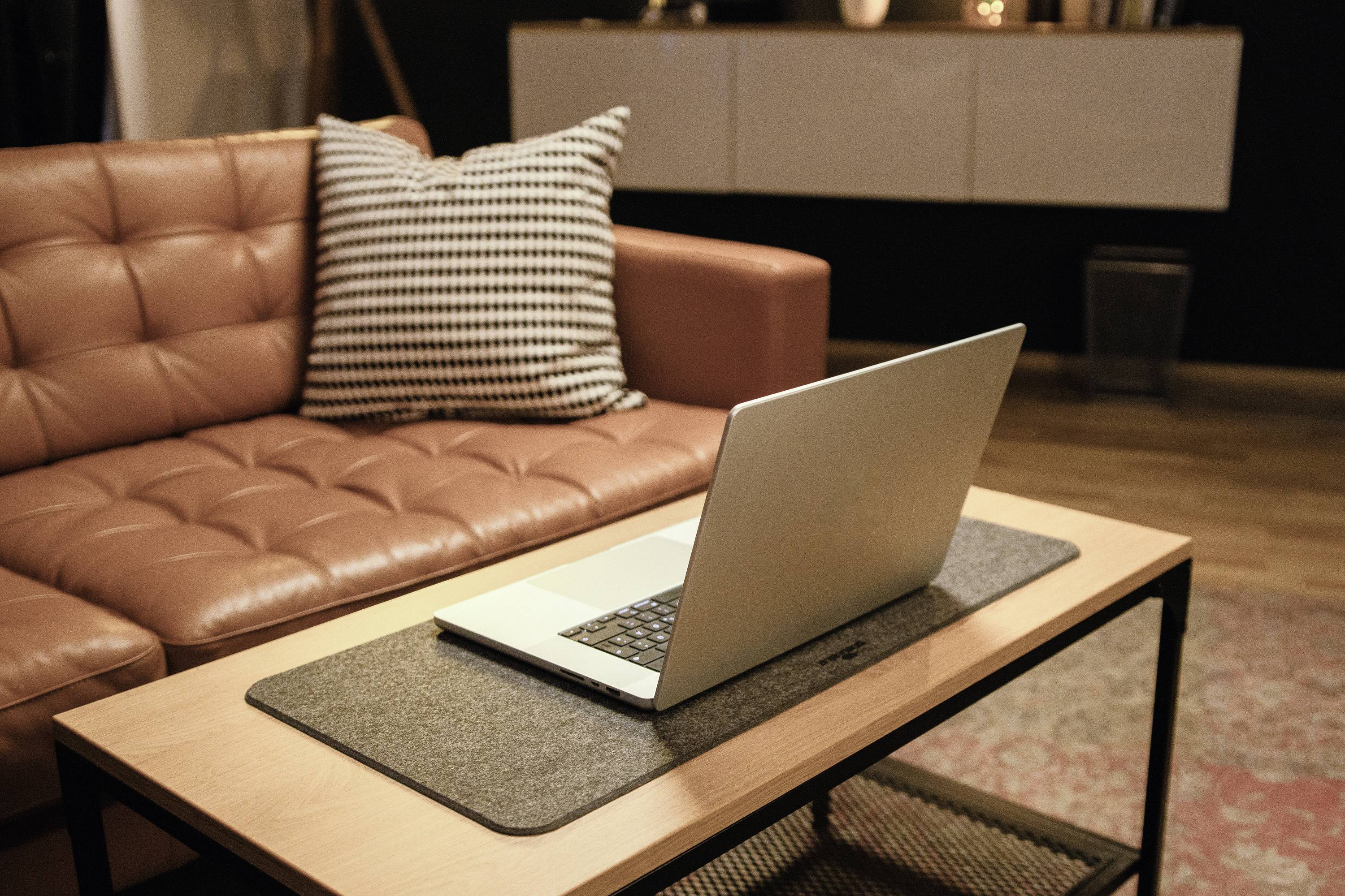 Ein Laptop steht auf einem Tisch im Wohnzimmer vor einem braunen Ledersofa mit einem schwarz-weiß karierten Kissen.