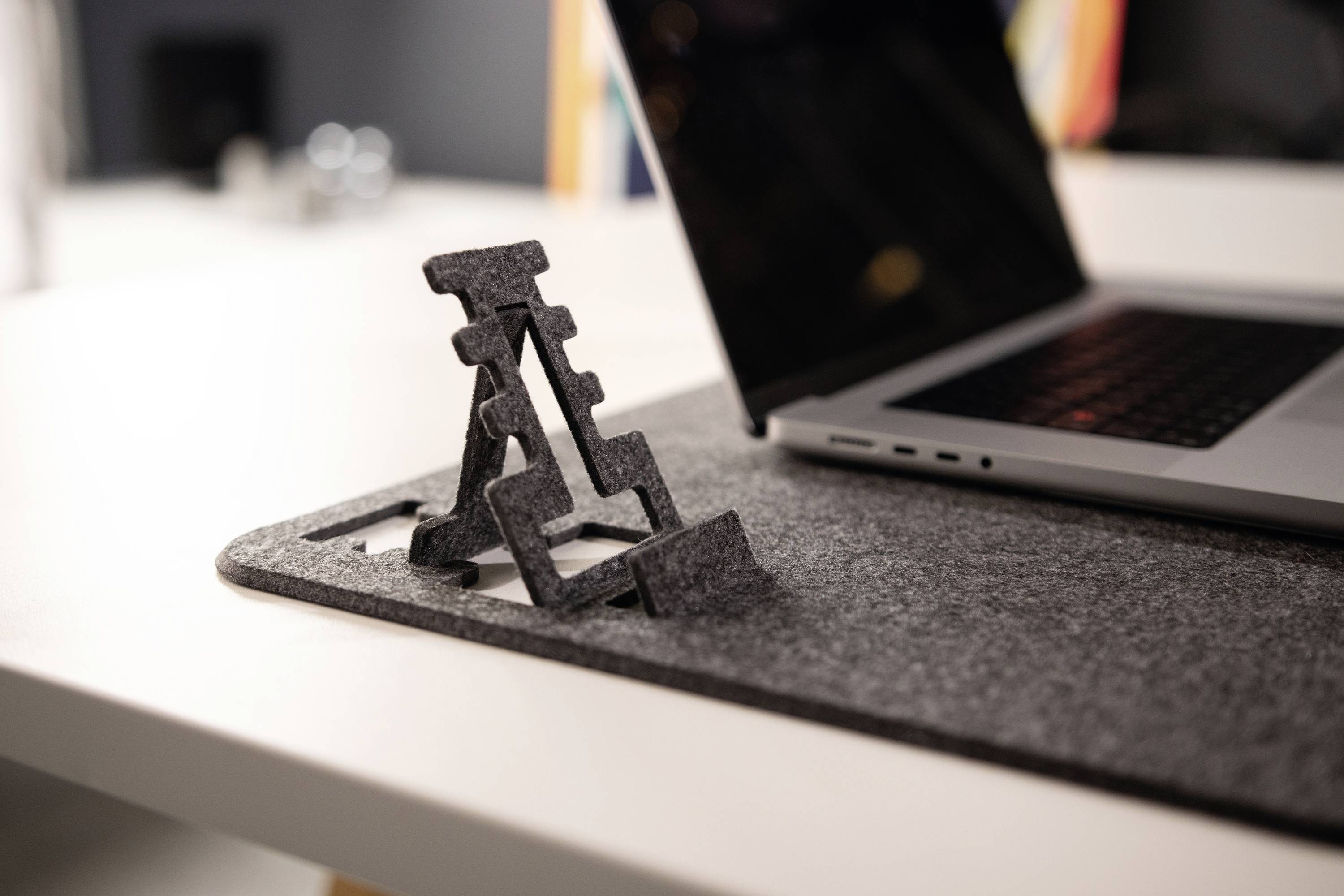 Ein Laptop steht auf einem Filzpad auf einem Tisch. Neben dem Laptop ist ein zusammenklappbarer Ständer aus Metall sichtbar.