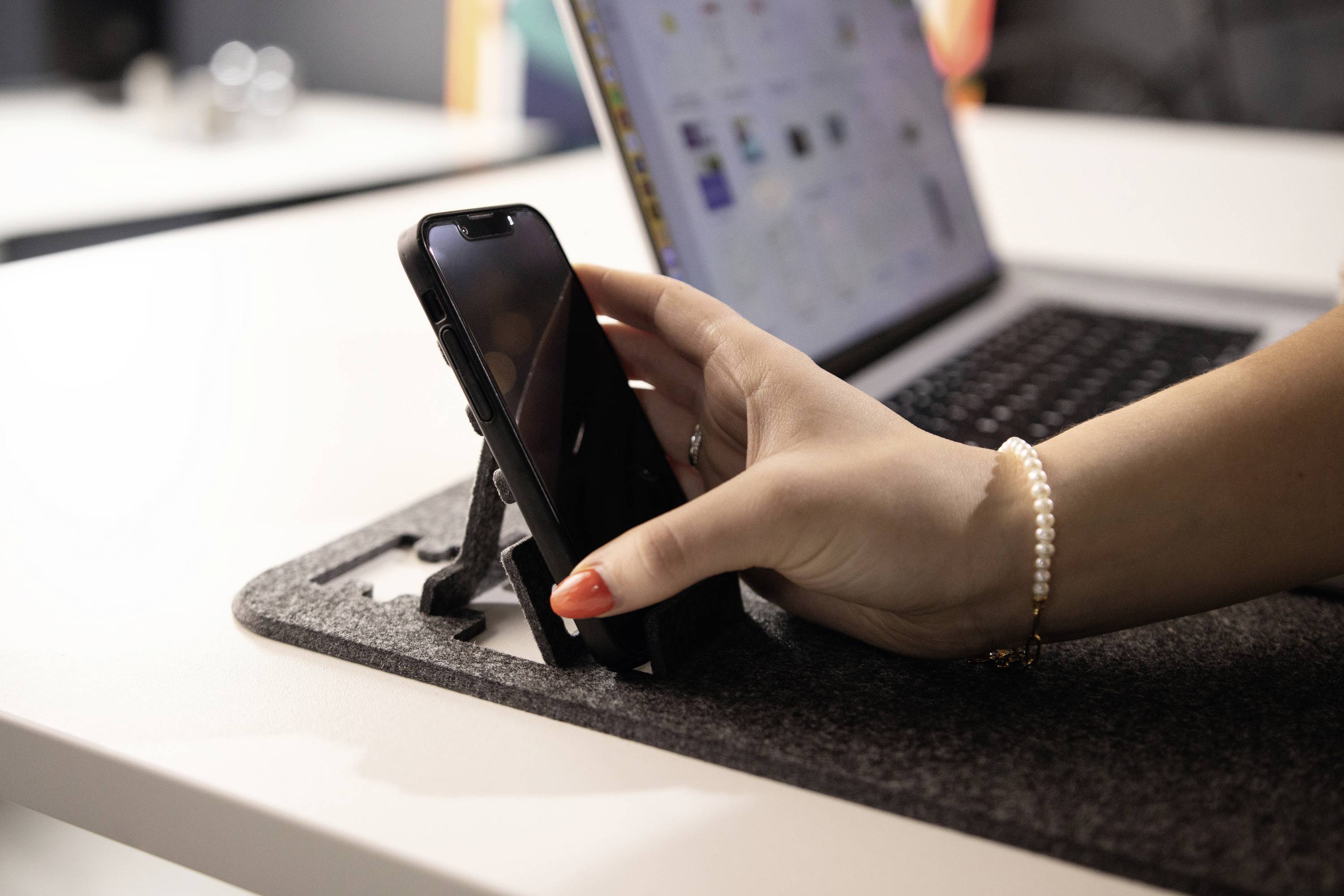 Eine Hand mit rotem Nagellack hält ein Handy auf einem Tisch neben einem geöffneten Laptop.