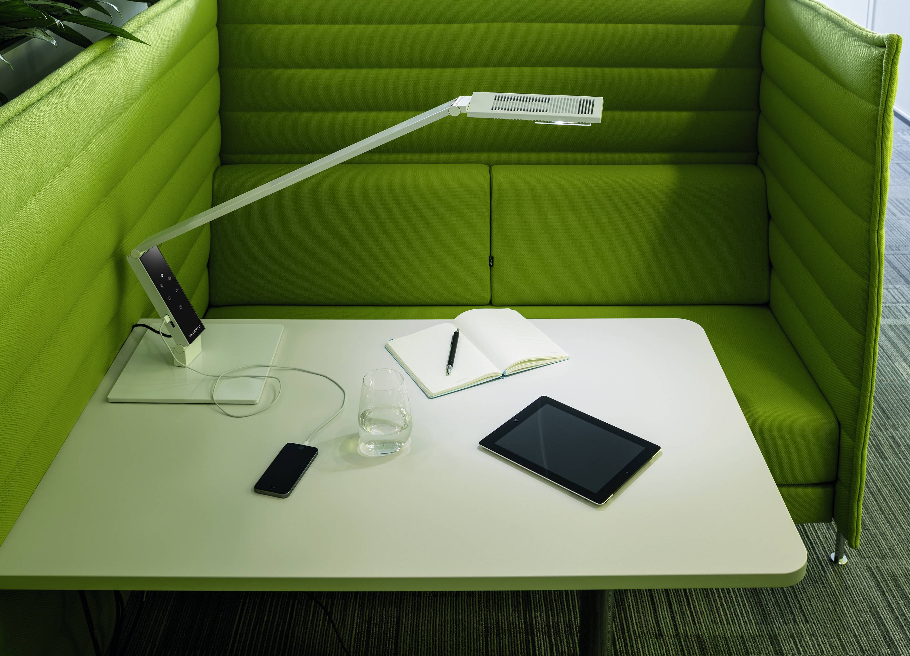 Runder Tisch mit einem Tablet, einem Notizbuch und einem Handy. Grüner gepolsterter Sitzbereich umgibt den Tisch auf drei Seiten.