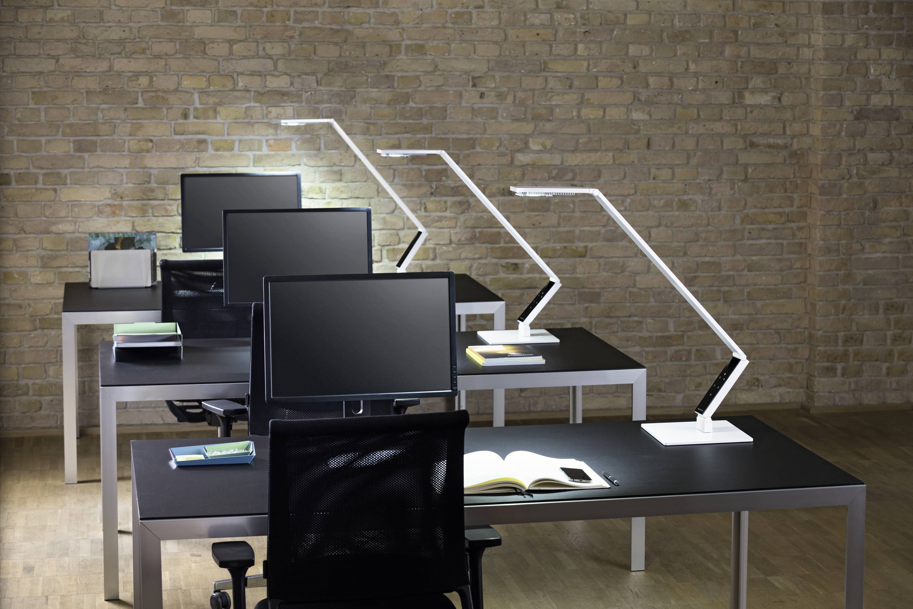 Ein modernes Büro mit vier Schreibtischen in einer Reihe. Jeder Tisch hat einen Monitor, eine Schreibtischlampe und ein offenes Buch.