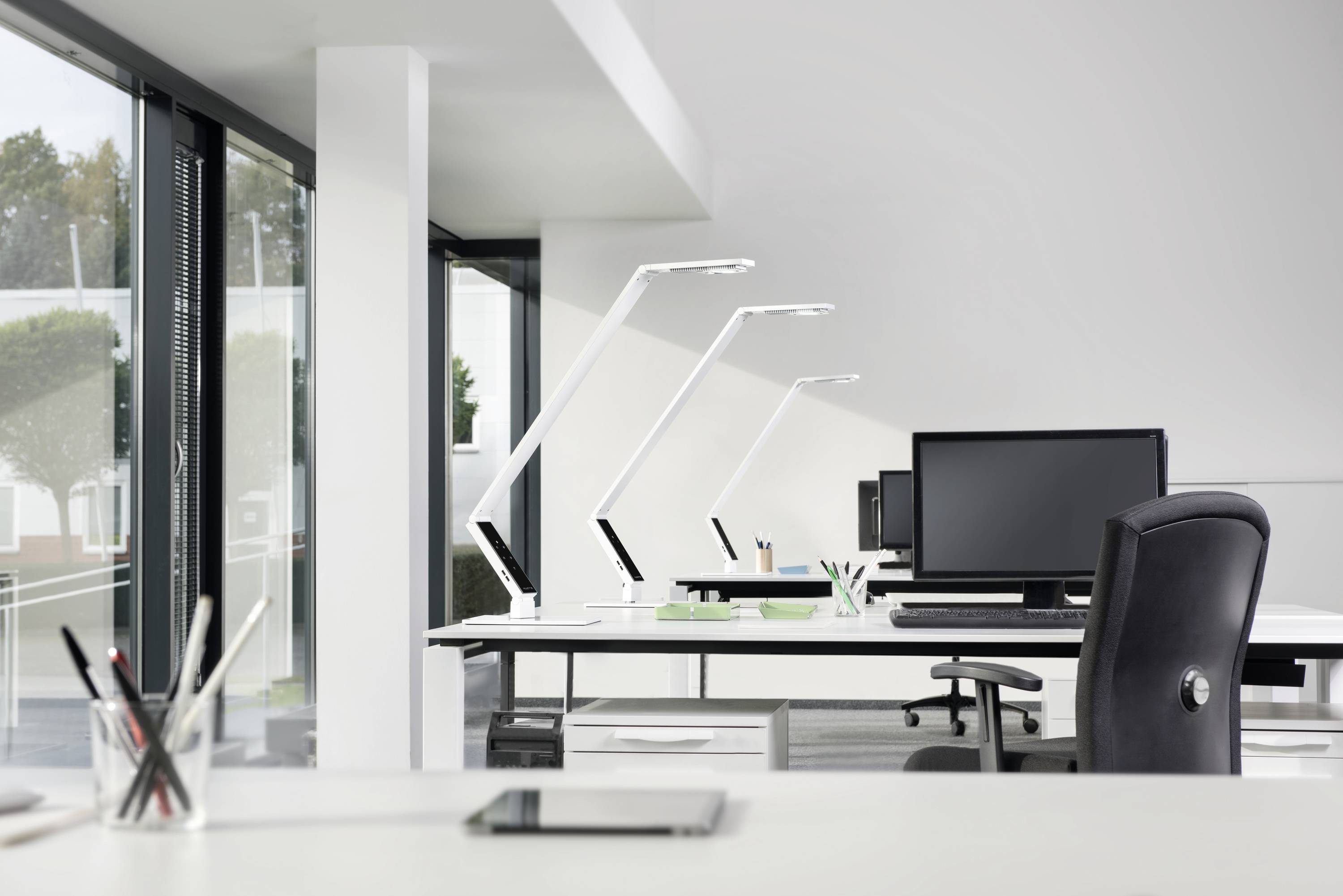 Ein modernes, helles Büro mit Schreibtischen, schwarzen Stühlen, Computermonitoren und Schreibtischlampen vor einer großen Fensterfront.