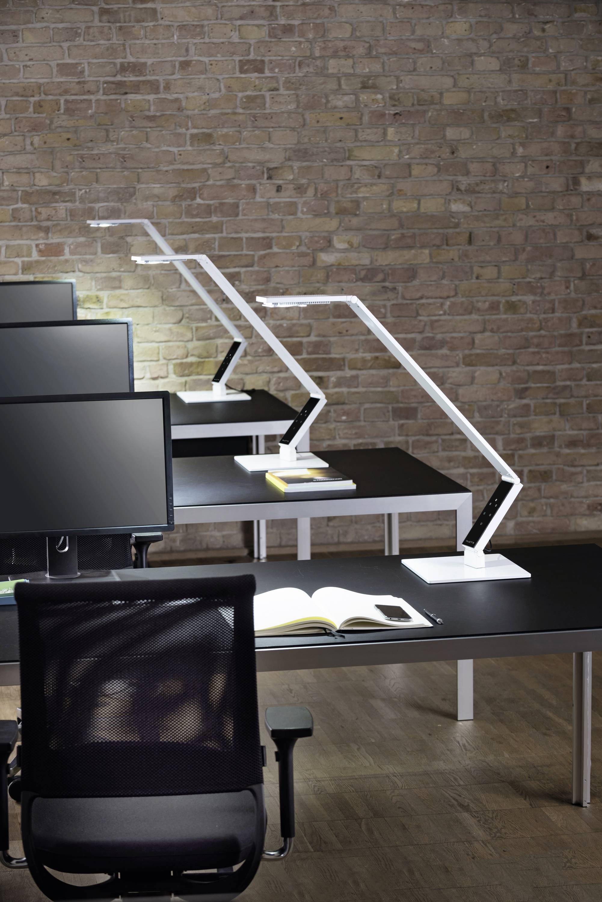 Moderne Büroeinrichtung mit schwarzen Schreibtischen, weißen Lampen und Monitoren vor einer Backsteinmauer. Ein offenes Buch liegt auf einem Tisch.
