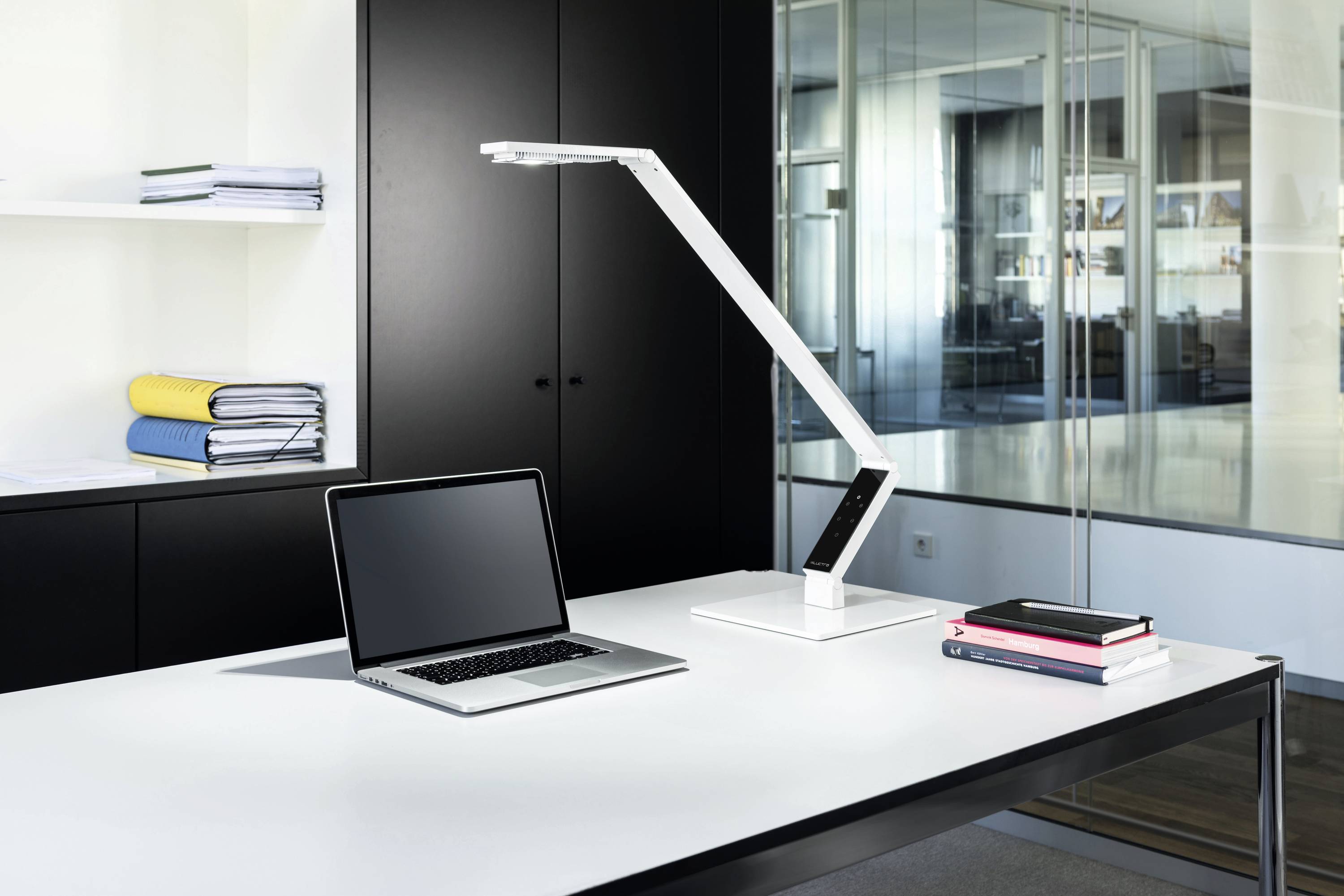 Ein moderner Schreibtisch in einem Büro mit einem Laptop, einer weißen Tischlampe und gestapelten Büchern auf einem weißen Tisch. Im Hintergrund sind Aktenschränke und Glaswände.