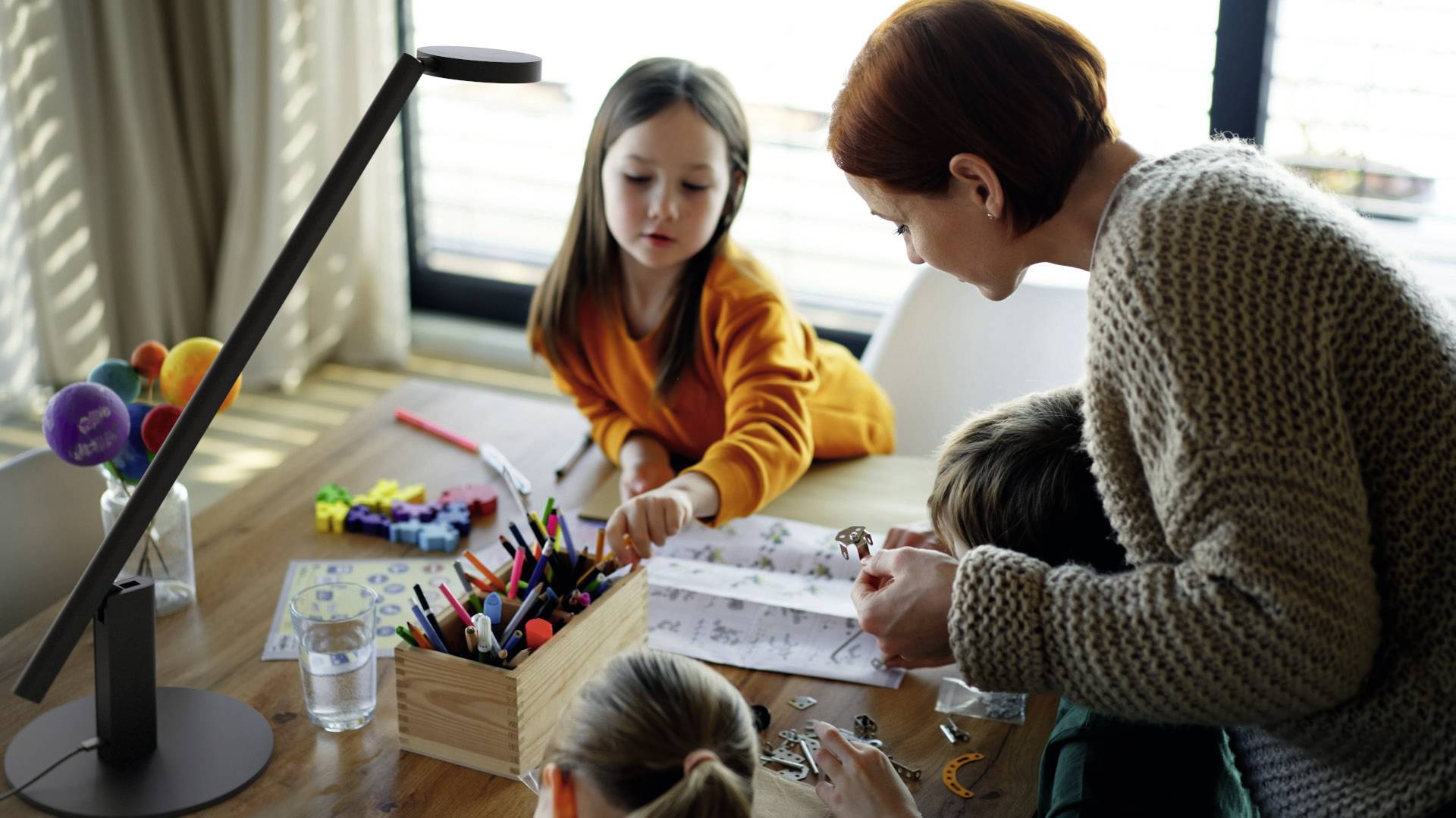 Eine Frau und drei Kinder sitzen an einem Tisch und basteln zusammen. Auf dem Tisch liegen Bastelmaterialien und ein Behälter mit Stiften.
