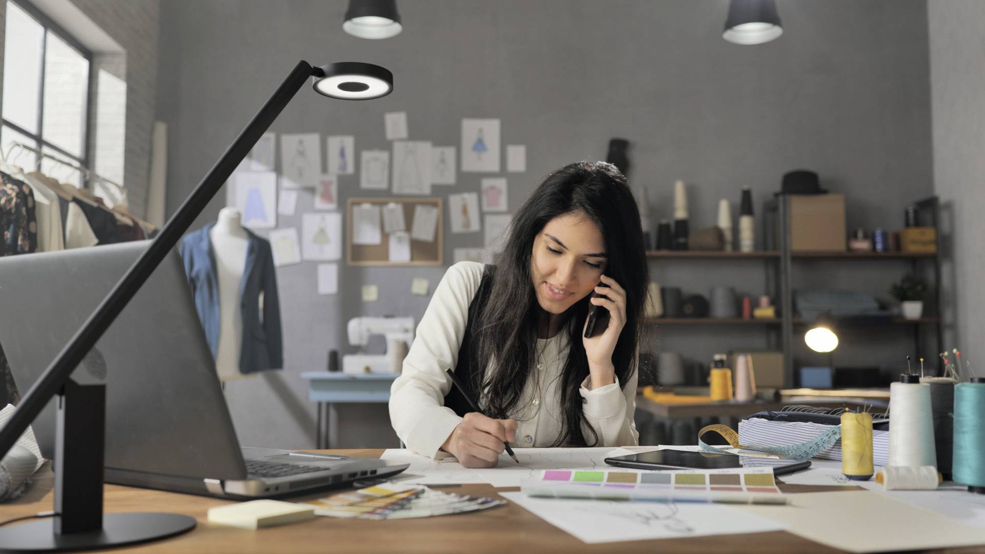 Eine Frau telefoniert in einem Designerbüro und macht Notizen. Im Hintergrund sind Stoffmuster und Modeentwürfe sichtbar.