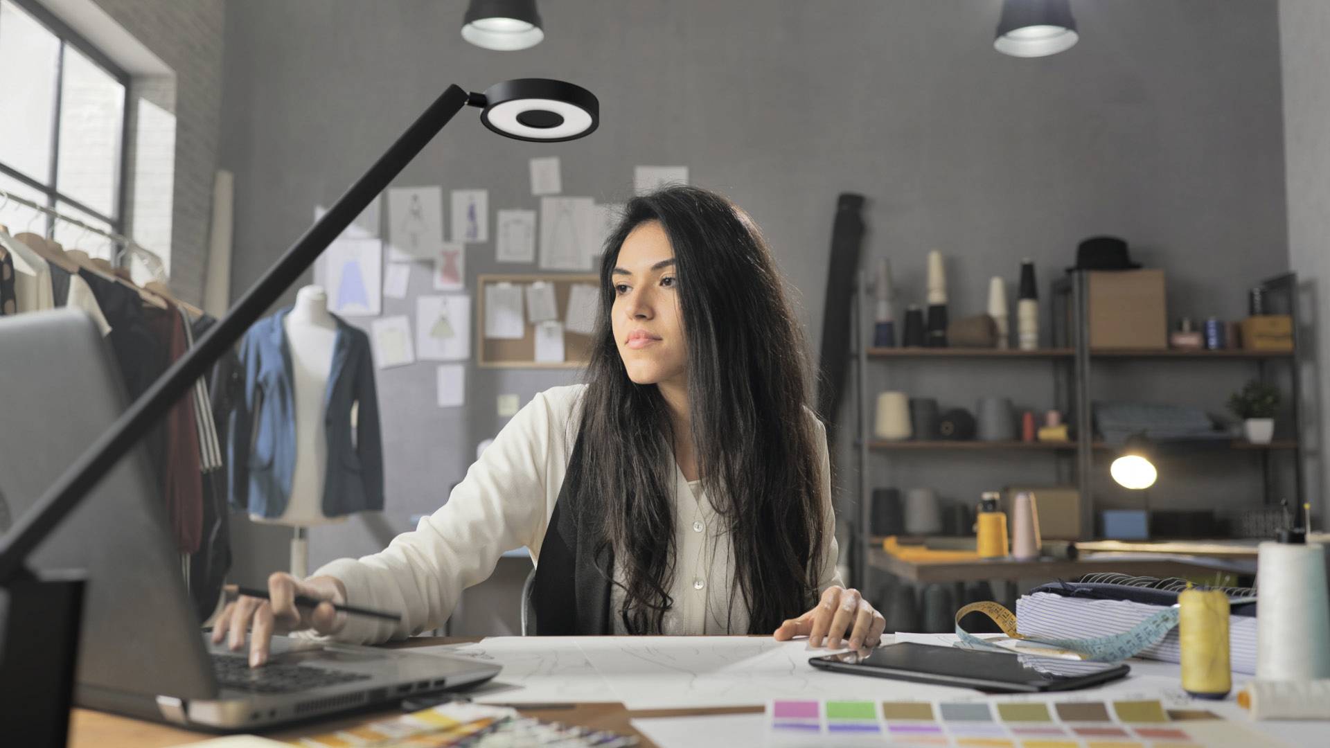 Junge Frau sitzt an einem Schreibtisch und arbeitet an einem Laptop in einem Modeatelier, umgeben von Textilien und Farbpaletten.