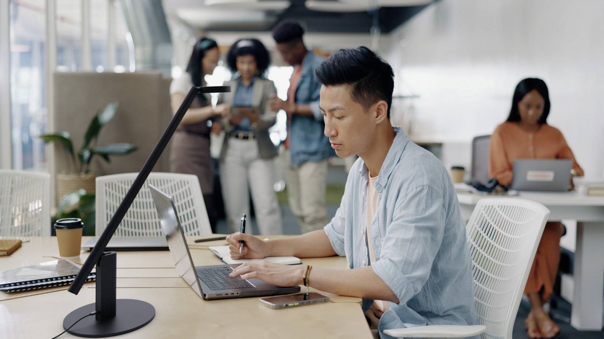 Ein konzentrierter Mann arbeitet an einem Laptop in einem modernen Großraumbüro. Im Hintergrund stehen Kolleg*innen zusammen und sprechen.