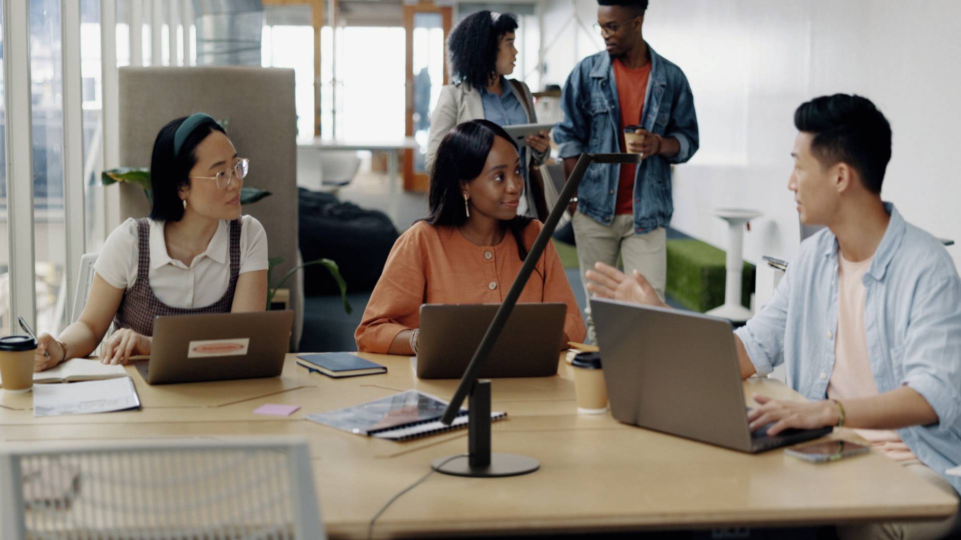 Drei Personen sitzen mit Laptops an einem Tisch und diskutieren angeregt. Zwei weitere Personen gehen im Hintergrund vorbei. Büroumgebung.