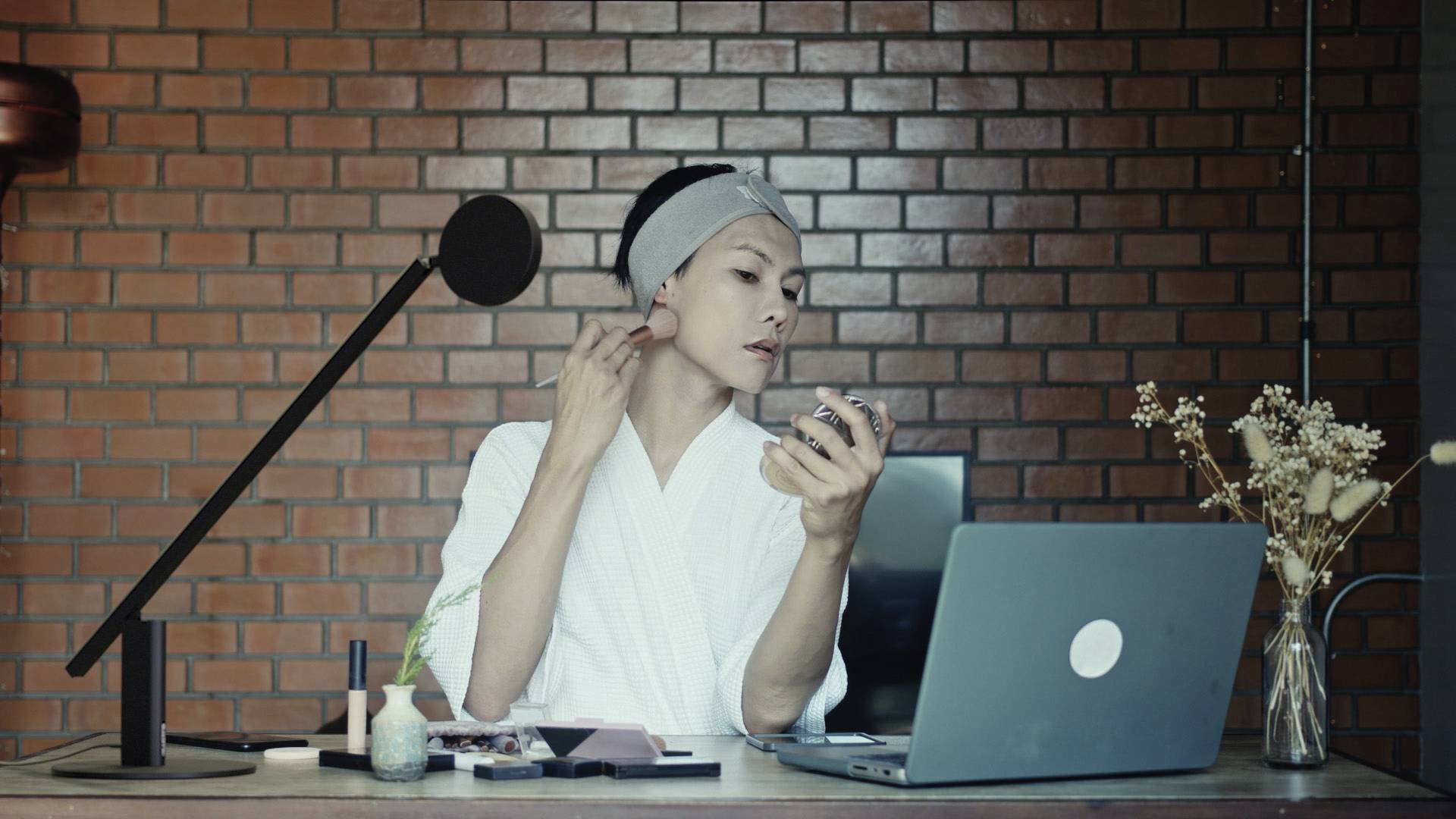 Eine Person in einem Bademantel trägt Make-up vor einem Laptop auf, mit einem Mikrofon auf dem Tisch.