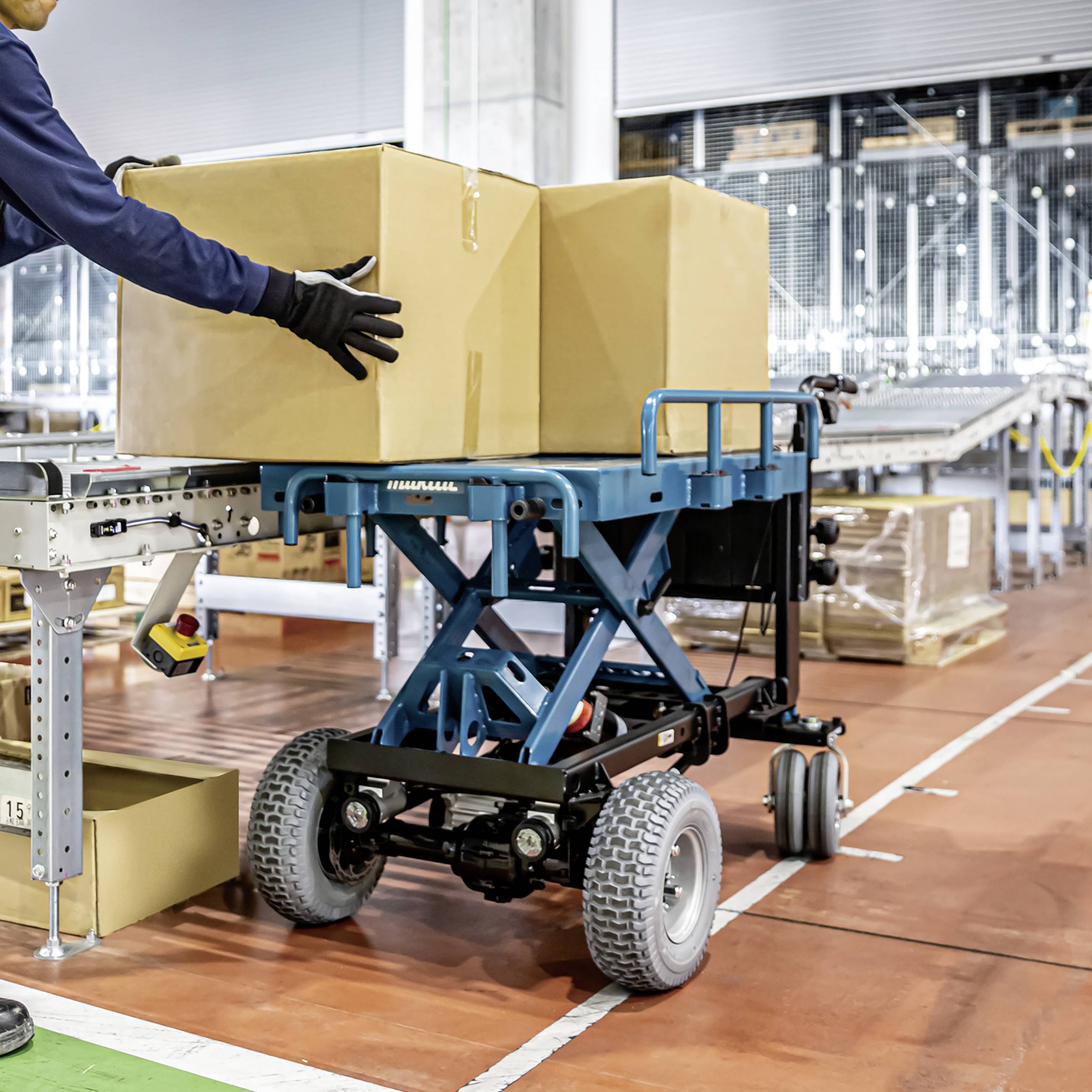 Ein Arbeiter schiebt einen hydraulischen Hubwagen mit großen Kartons in einem Lager. Im Hintergrund sind Regale und weitere Kartons zu sehen.
