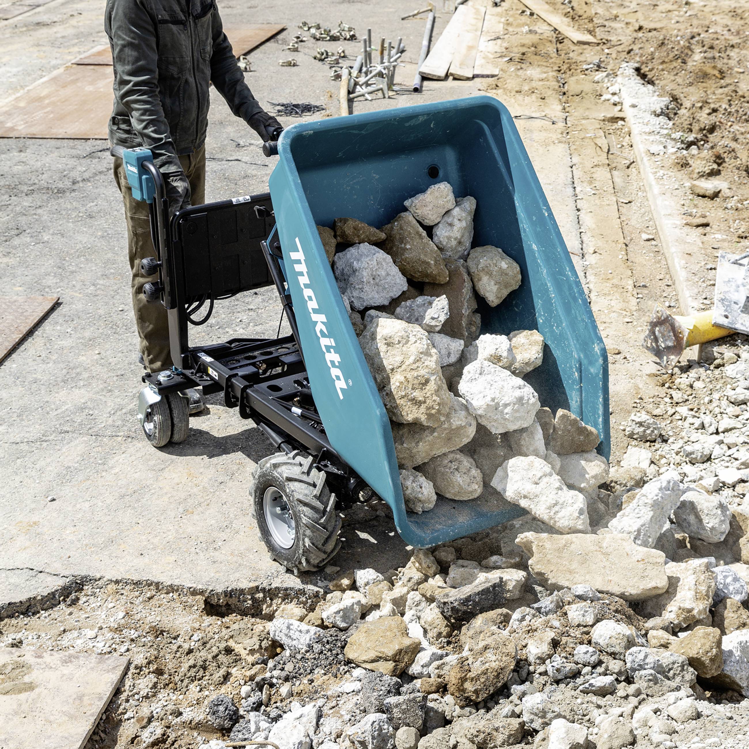 Eine Person bedient eine elektrische Schubkarre mit großen Steinen auf einer Baustelle. Die Konstruktion im Hintergrund deutet auf Bauarbeiten hin.