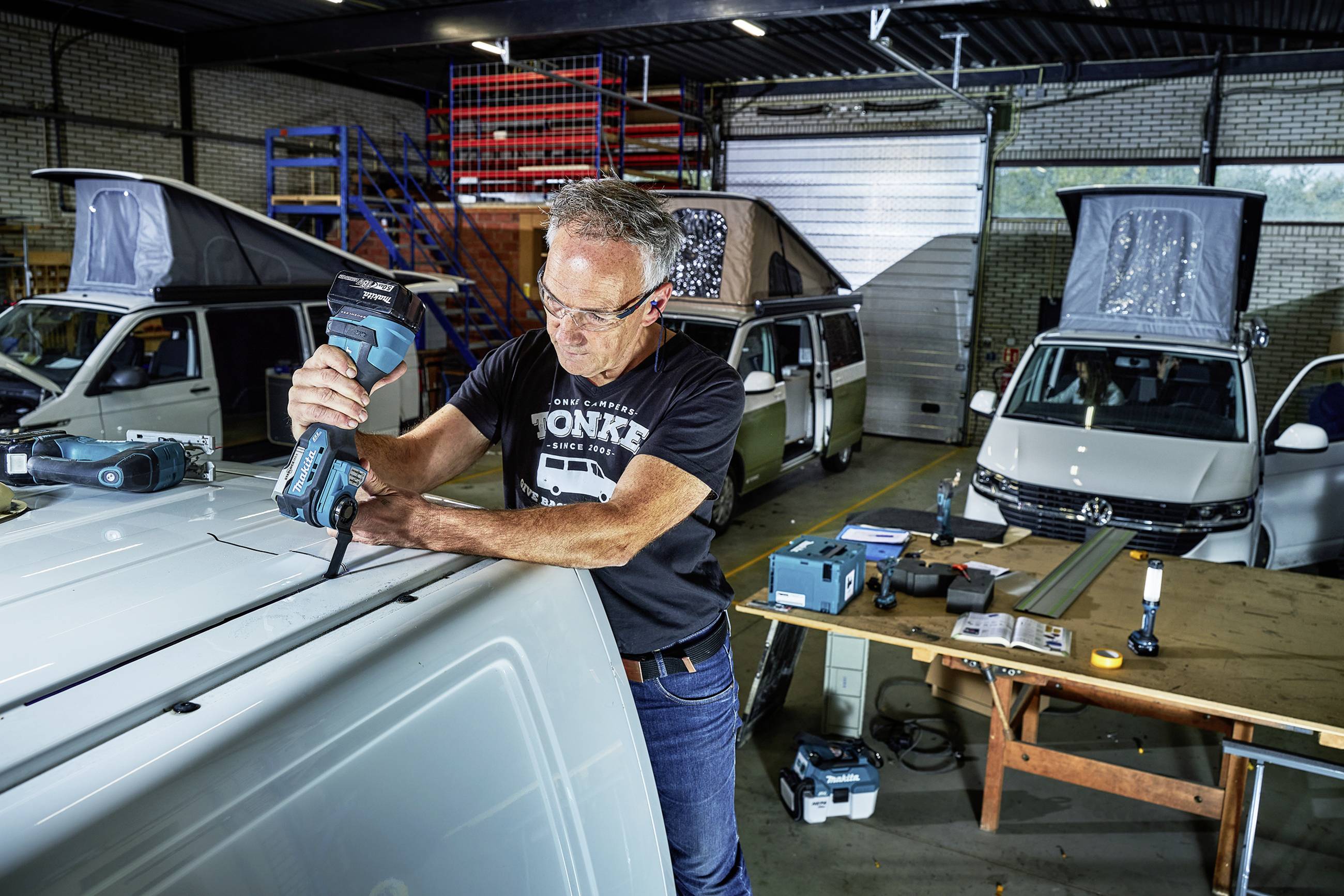 Ein Mann mit Schutzbrille arbeitet in einer Werkstatt an einem Transporter mit einem Elektrowerkzeug. Im Hintergrund sind weitere Fahrzeuge zu sehen.