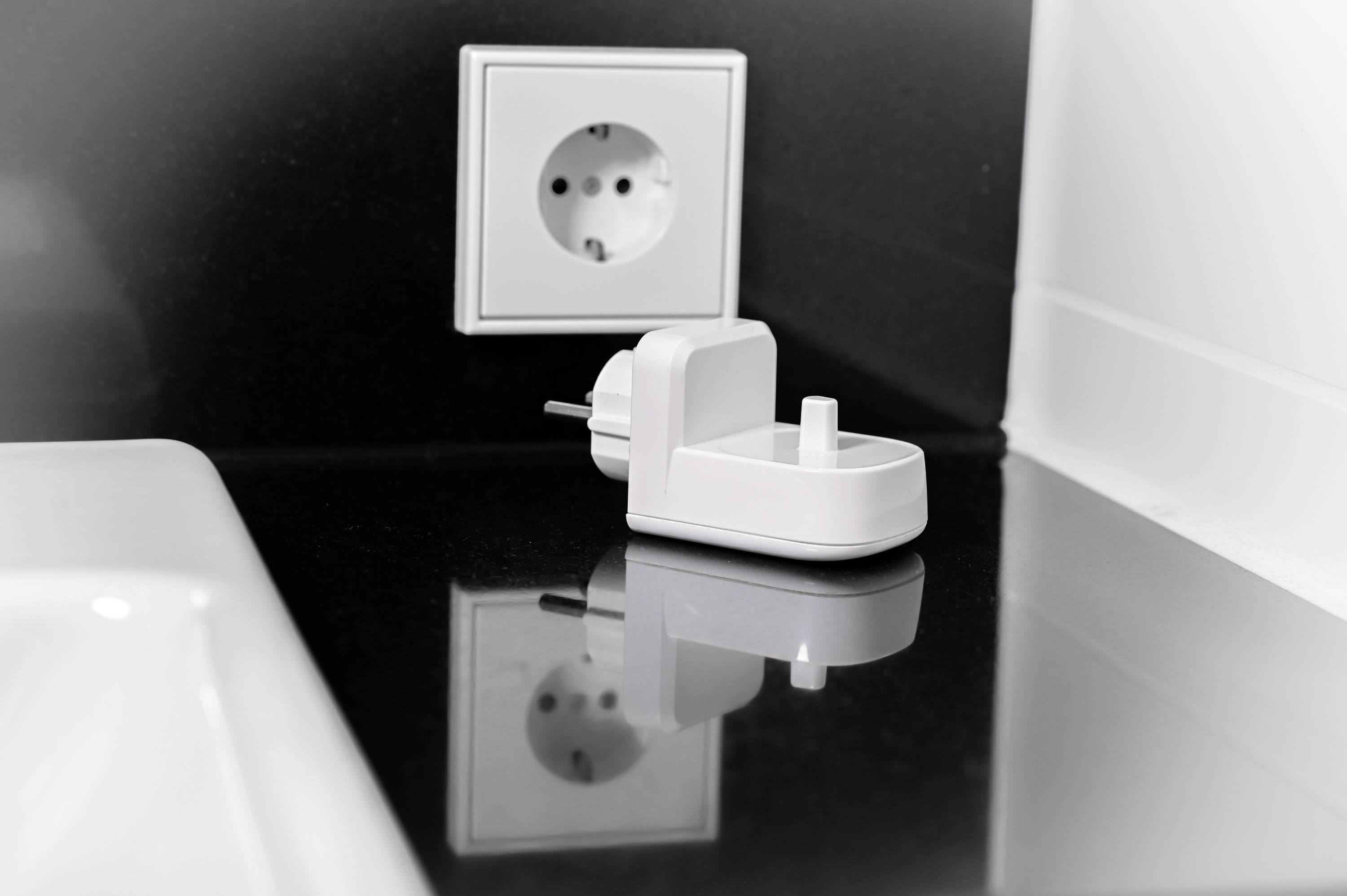 Eine weiße Steckdose an einer schwarzen Wand mit einem weißen Netzstecker auf der Arbeitsfläche. Es gibt ein Waschbecken daneben.