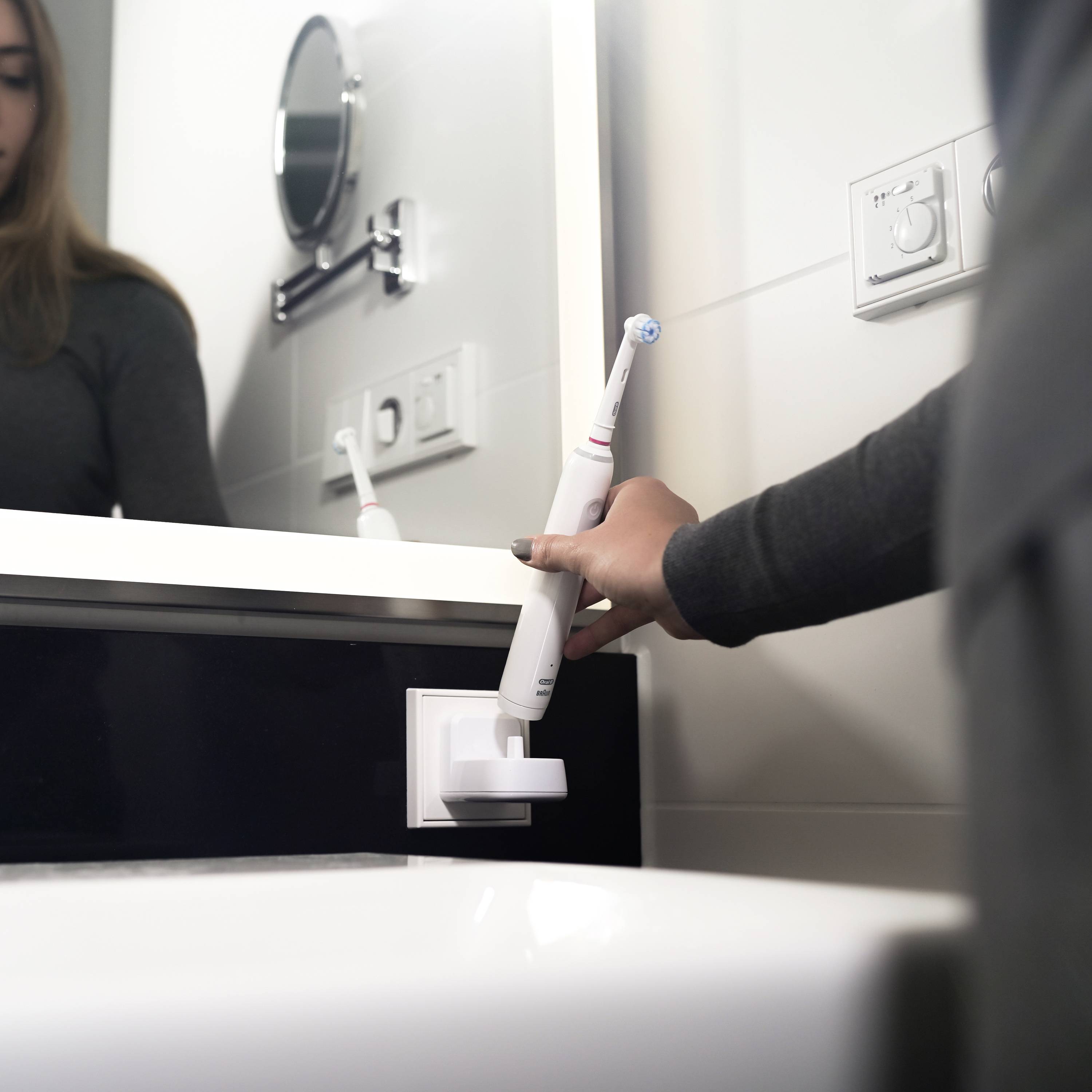 Eine Person lädt eine elektrische Zahnbürste an einer Steckdose im Badezimmer auf. Im Hintergrund sind ein Spiegel und Schalter.