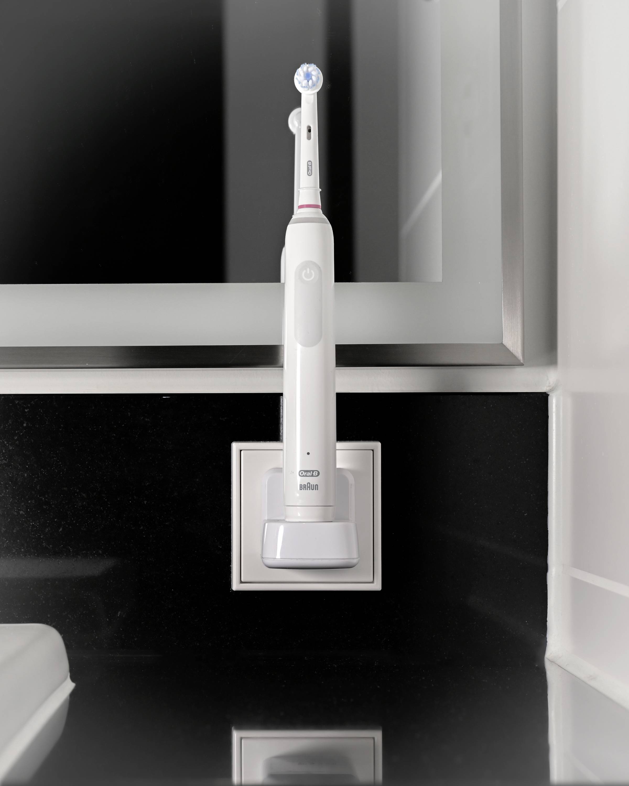 Elektrische Zahnbürste in Ladestation auf schwarzer Ablage im Bad, vor silbernem Spiegel montiert.