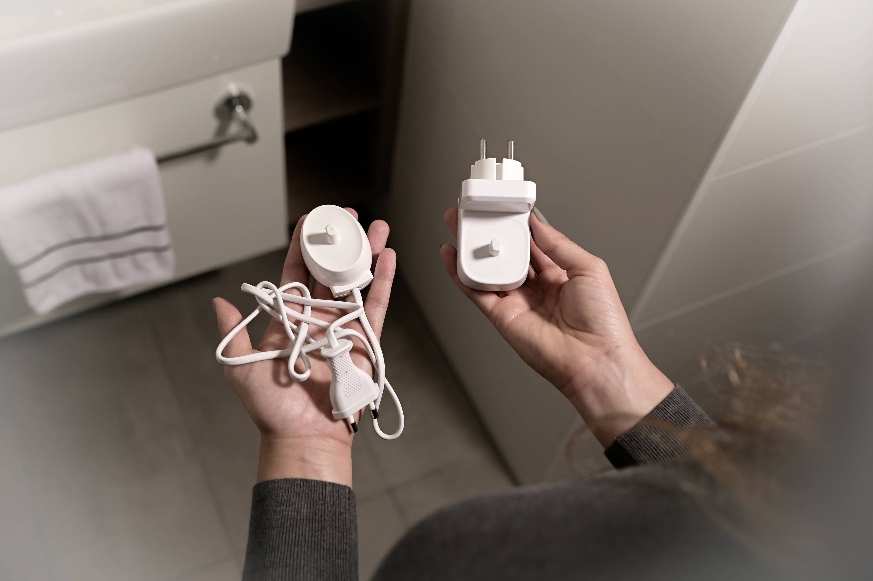 Eine Person hält ein weißes Rasier-Steckdosenladegerät und ein loses Kabel in den Händen, stehend im Badezimmer.