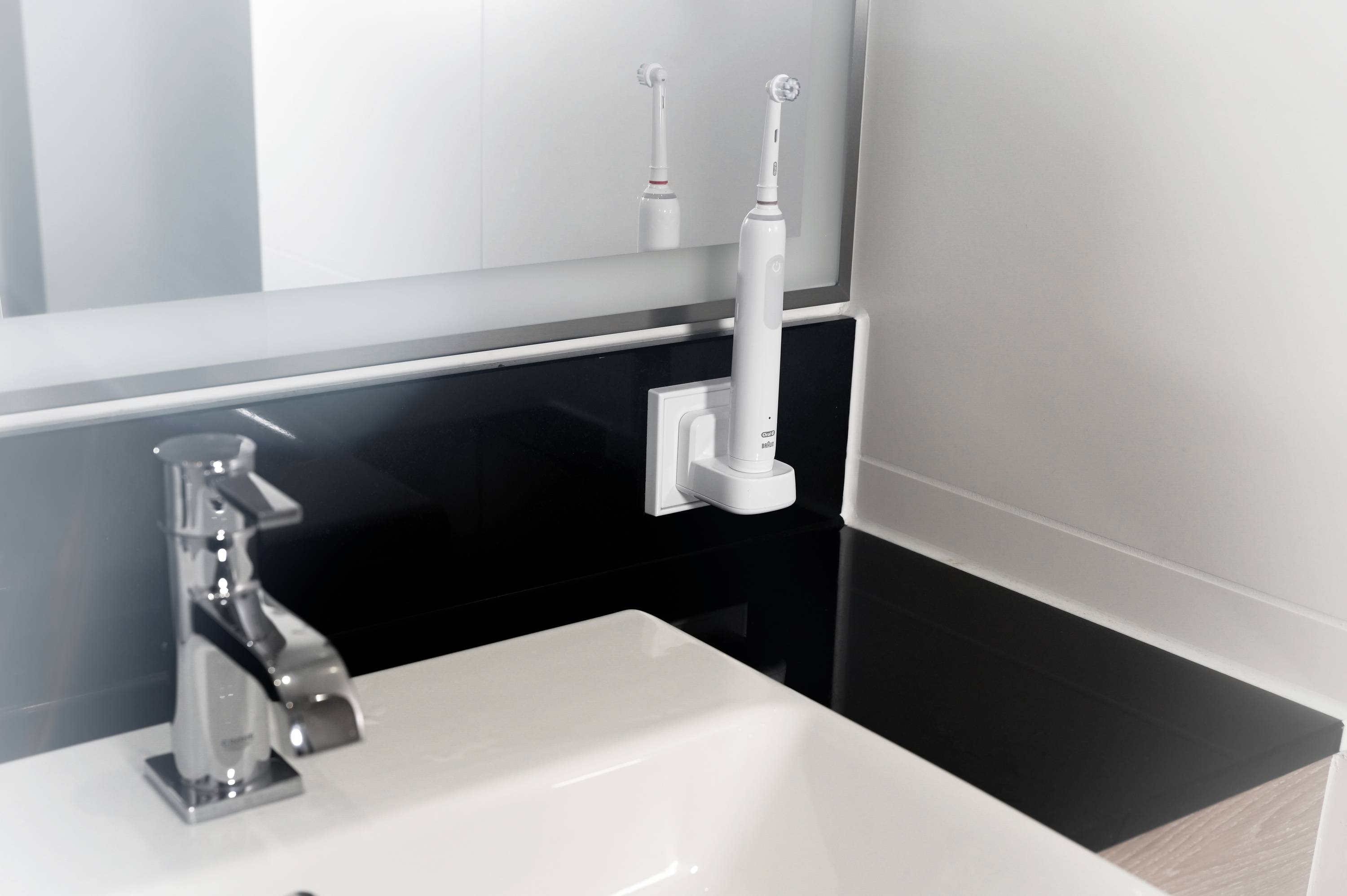 Ein Badezimmer mit weißem Waschbecken und Wasserhahn, daneben eine elektrische Zahnbürste in einer Halterung an der Wand.