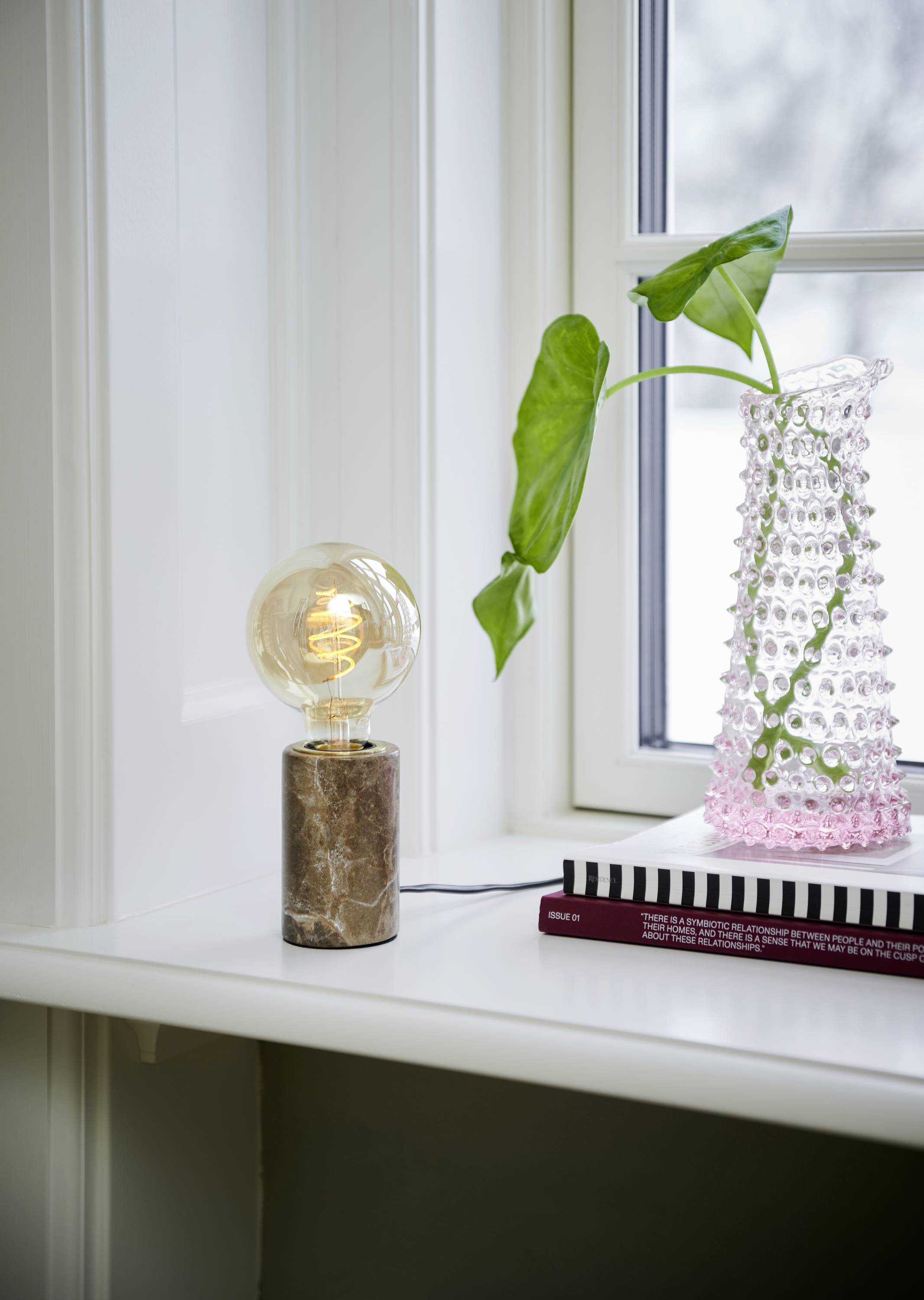 Eine moderne Glühlampe mit Marmorbasis steht neben einem rosa, strukturierten Glasgefäß mit grünem Blatt auf einer Fensterbank.