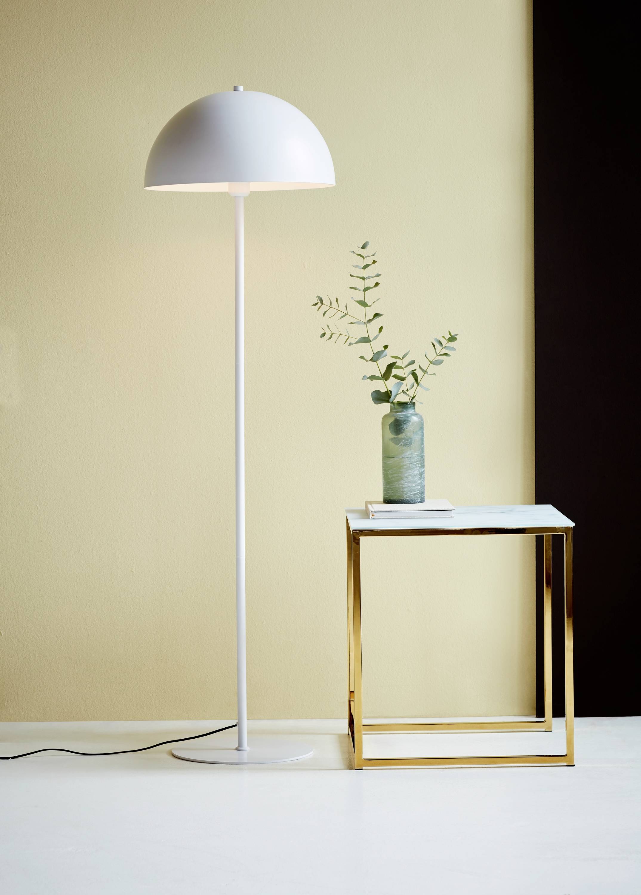 Stehlampe mit weißem Schirm neben einem kleinen Tisch mit goldenen Beinen und einer Vase mit Grünpflanze, vor hellgelber Wand.