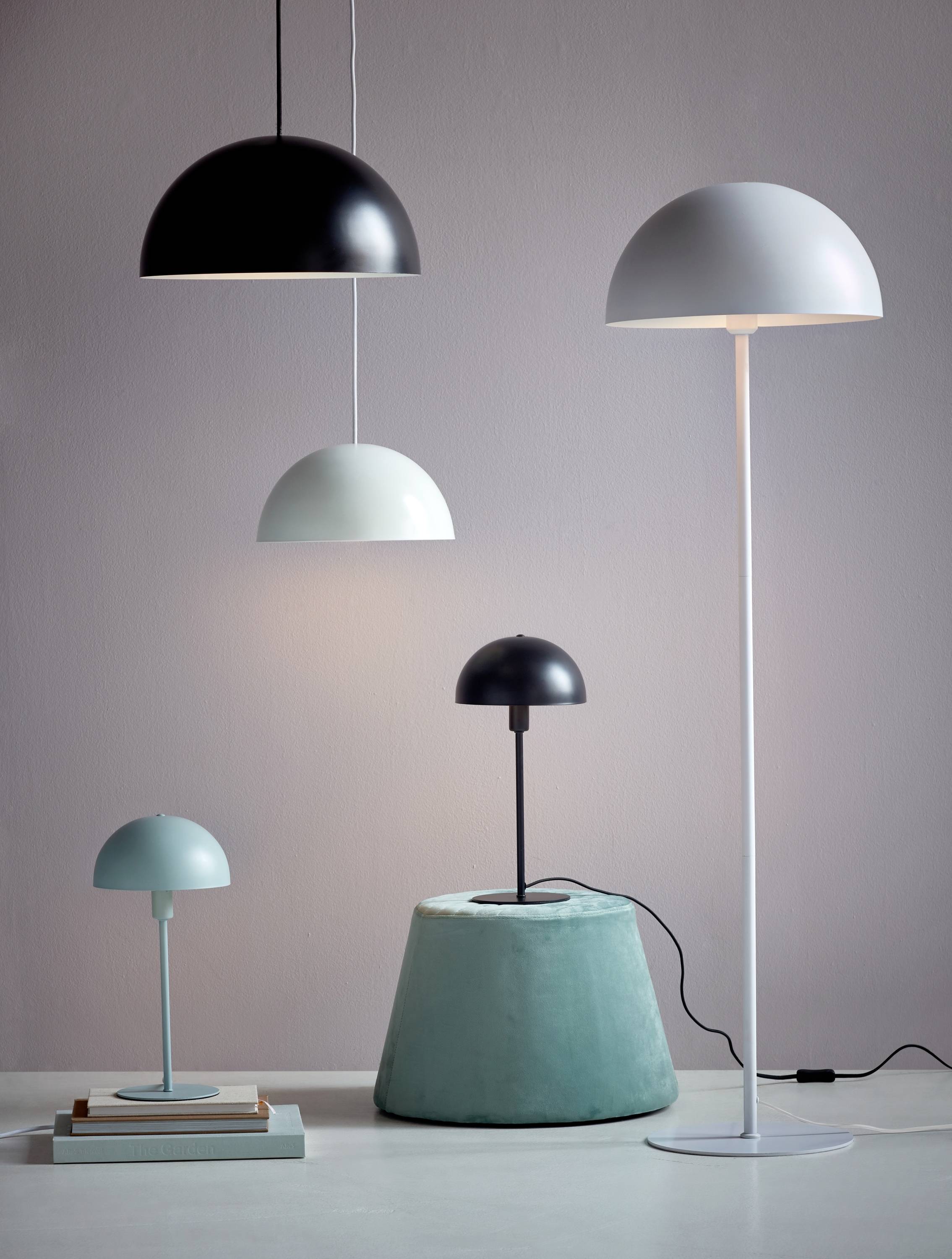 Vier moderne Lampen in verschiedenen Farben und Größen stehen auf einem Tisch und einem Podest vor einer hellgrauen Wand.