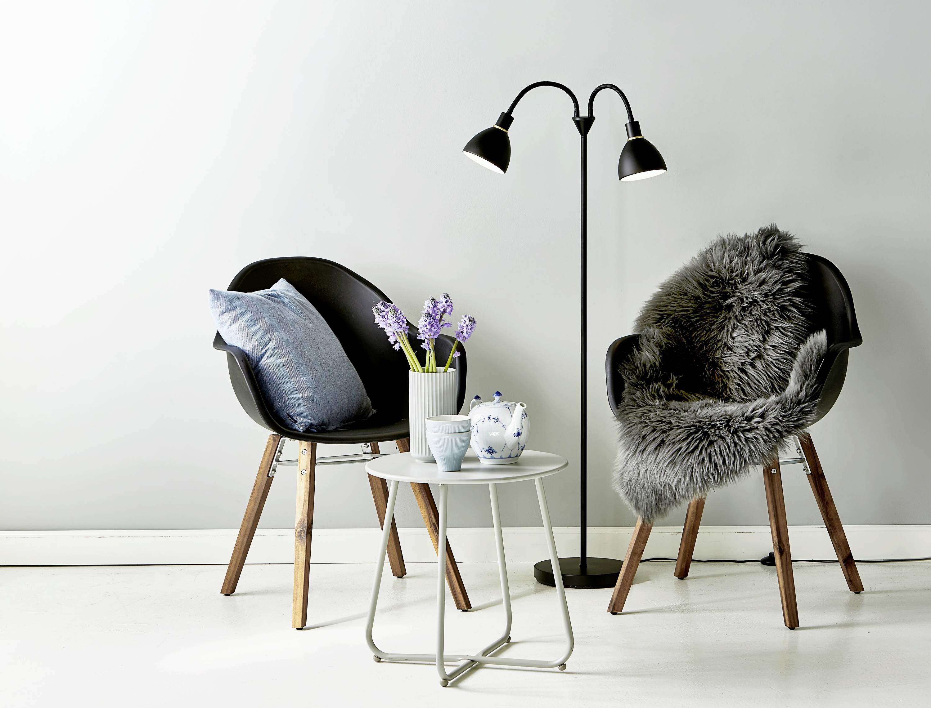 Zwei schwarze Stühle mit Holzkufen, einer mit grauem Fell, der andere mit blauem Kissen. Dazwischen ein kleiner Tisch mit Blumen und Teekanne. Im Hintergrund eine Stehlampe.