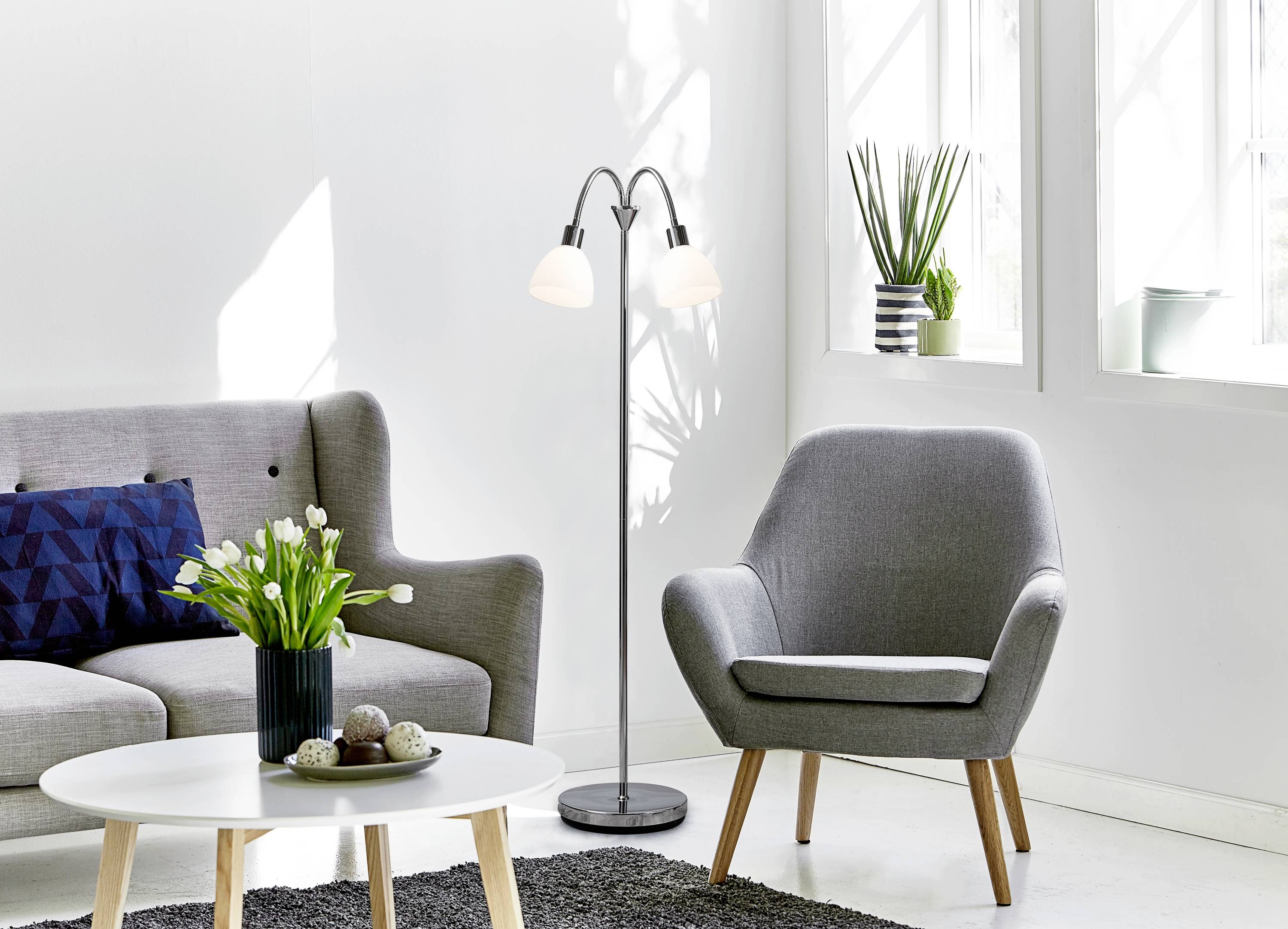 Helles Wohnzimmer im skandinavischen Stil mit grauem Sofa und Sessel, Stehlampe, weißen Wänden, Tisch, Teppich und Pflanzen.