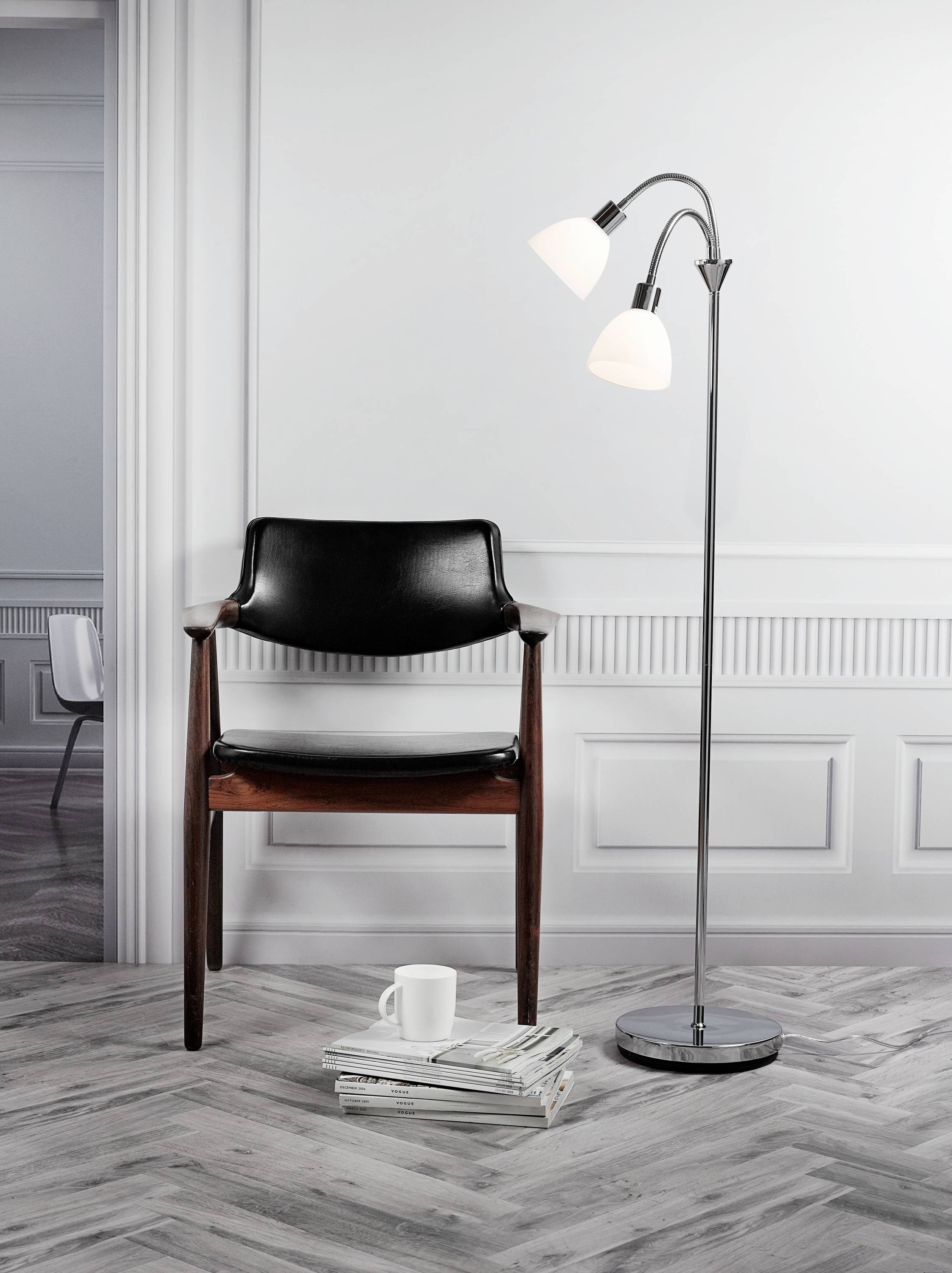 Schwarzer Stuhl und Stehlampe in einem eleganten Raum mit Parkettboden, neben einem Stapel Zeitschriften und einer weißen Tasse.