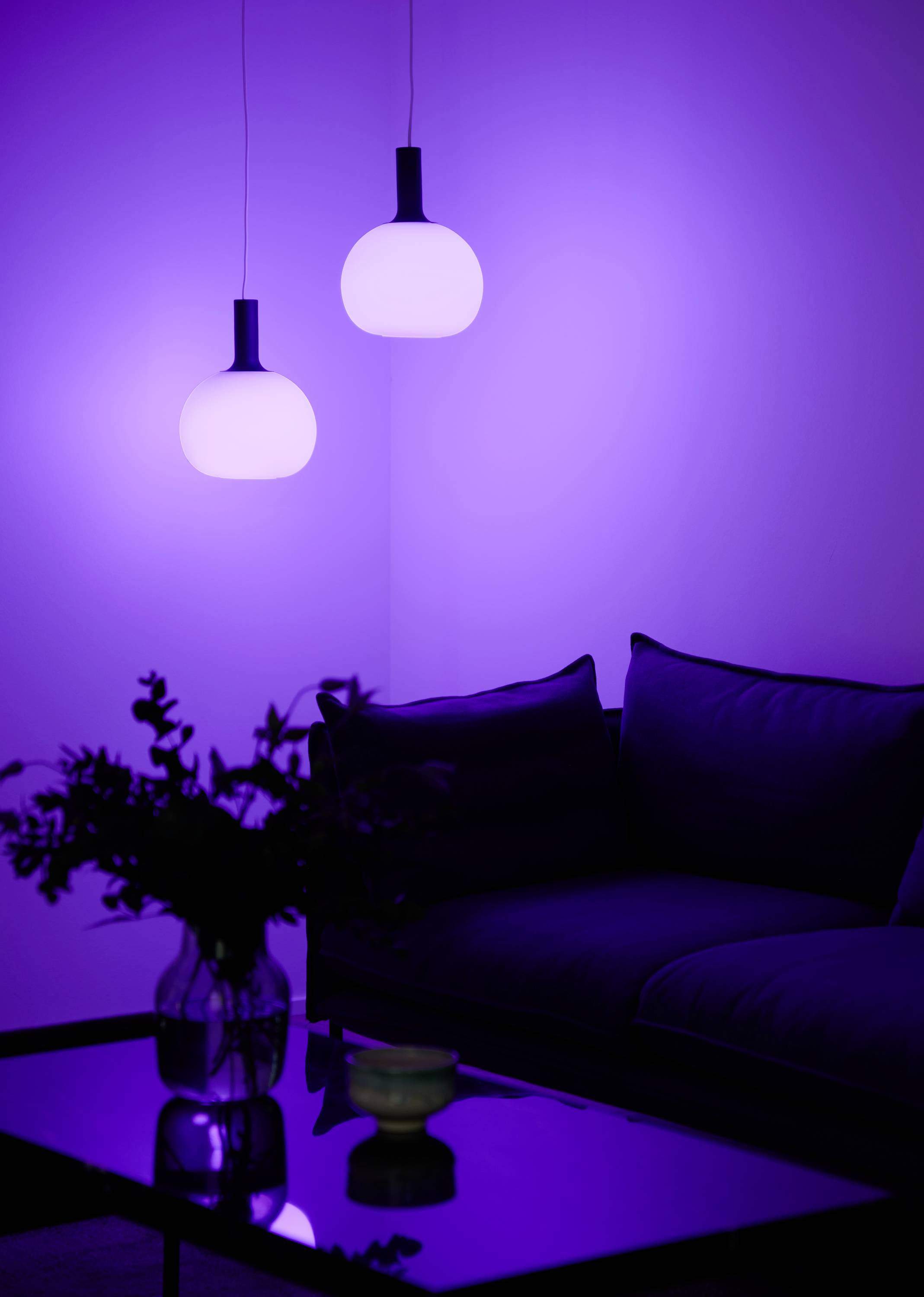 Zwei leuchtende Hängeleuchten in einer modernen Einrichtung, die einen violetten Schein auf ein Sofa und einen Glastisch werfen.