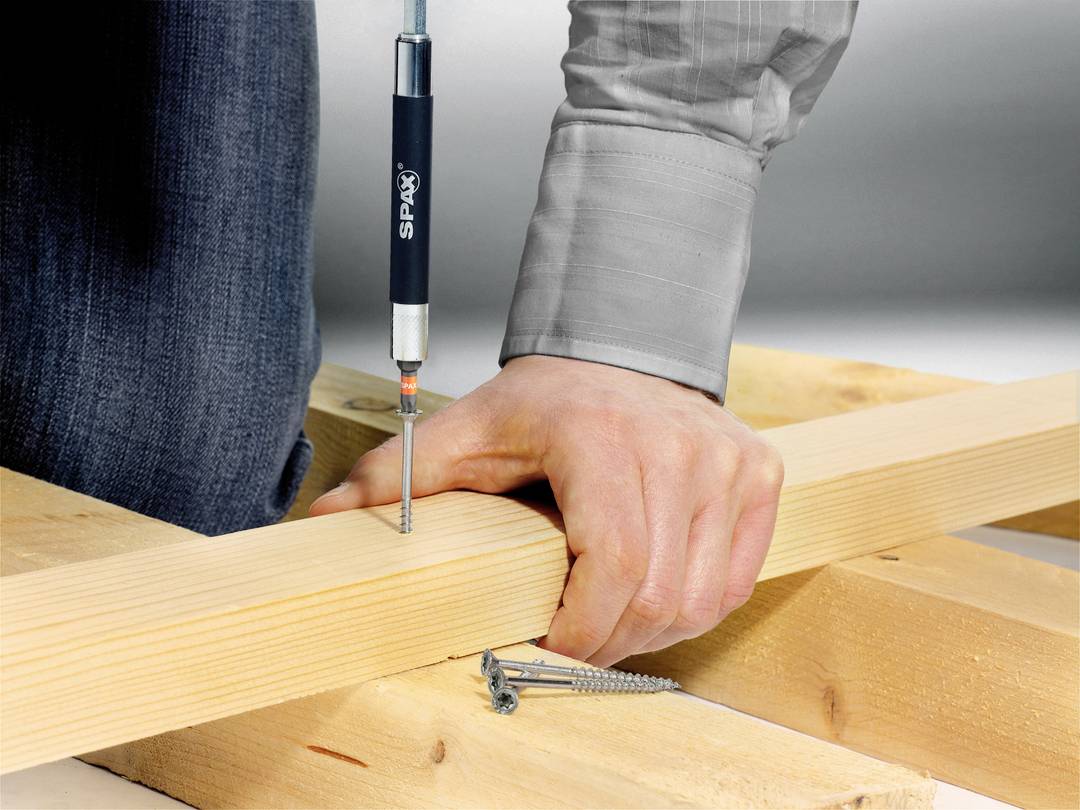 Eine Person schraubt mit einem Schraubenzieher eine Schraube in ein Holzbrett. Mehrere Schrauben liegen daneben.