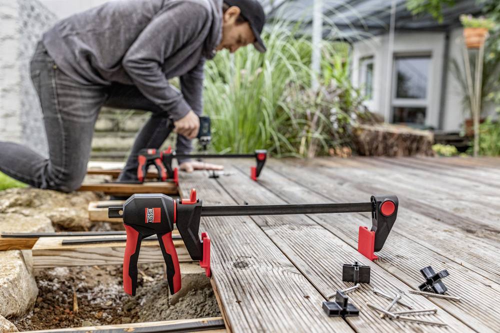 Eine Person baut eine Terrasse und verwendet rote und schwarze Zwingen, um Holzdielen zu befestigen. Schrauben und Werkzeuge liegen bereit.