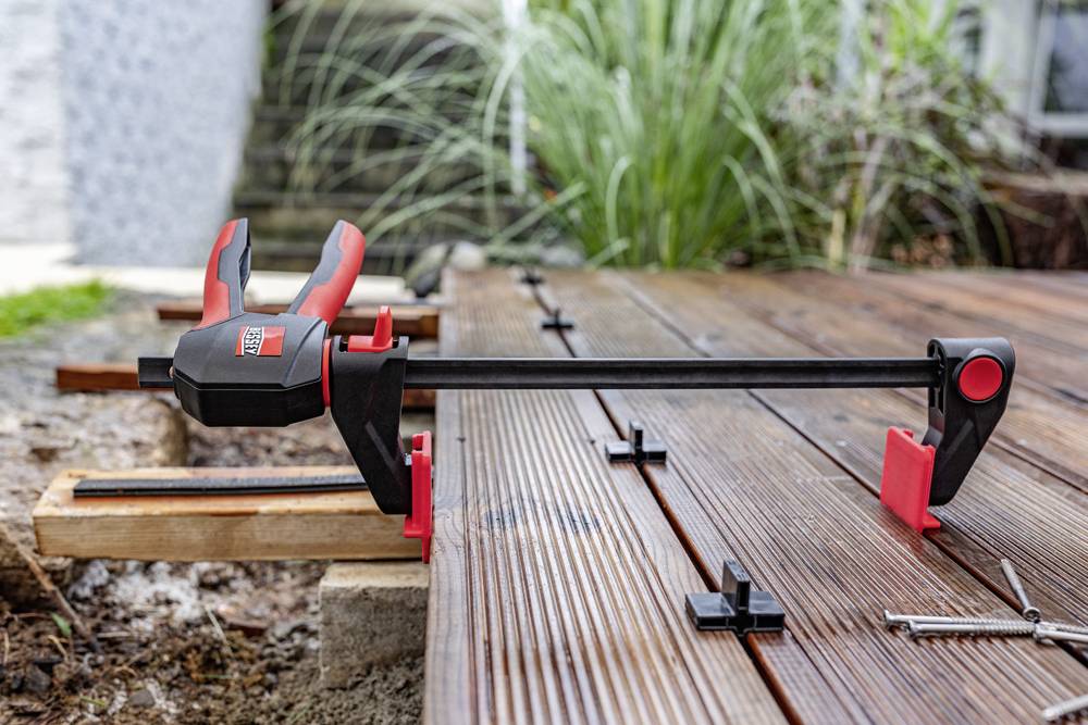 Eine Zwinge hält Holzbretter auf einer Terrasse zusammen. Im Hintergrund sind unscharfe Pflanzen und weitere Bretter zu sehen.
