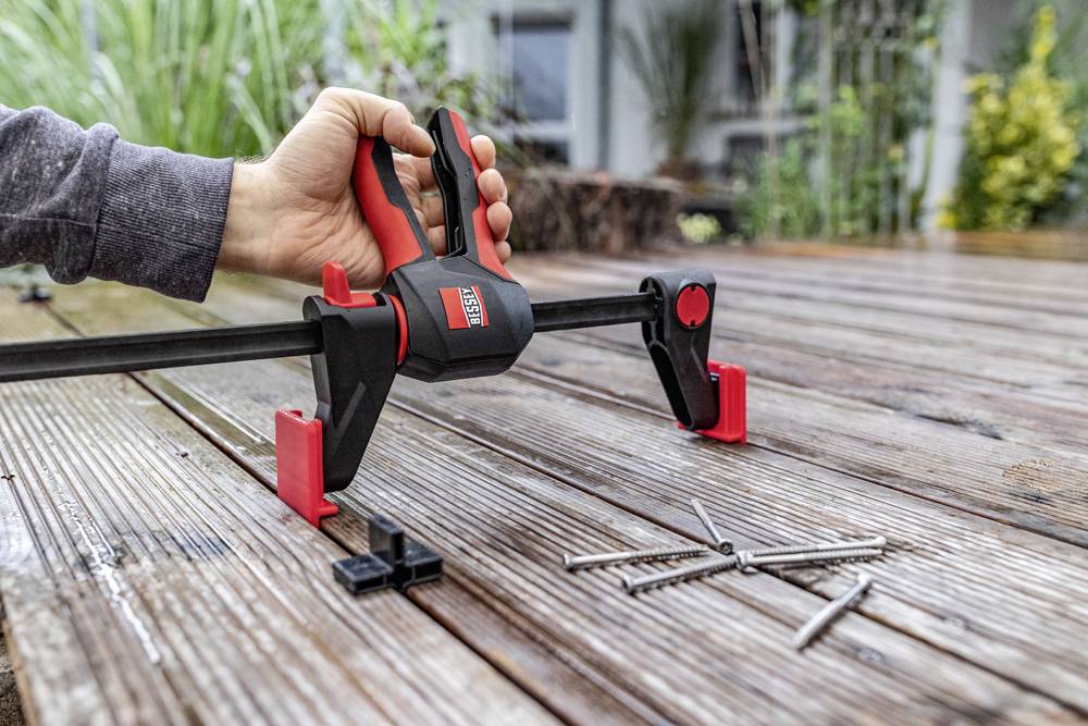 Eine Hand benutzt eine rote Schraubzwinge auf einem Holztisch im Freien. Einige Schrauben liegen daneben. Im Hintergrund Pflanzen.