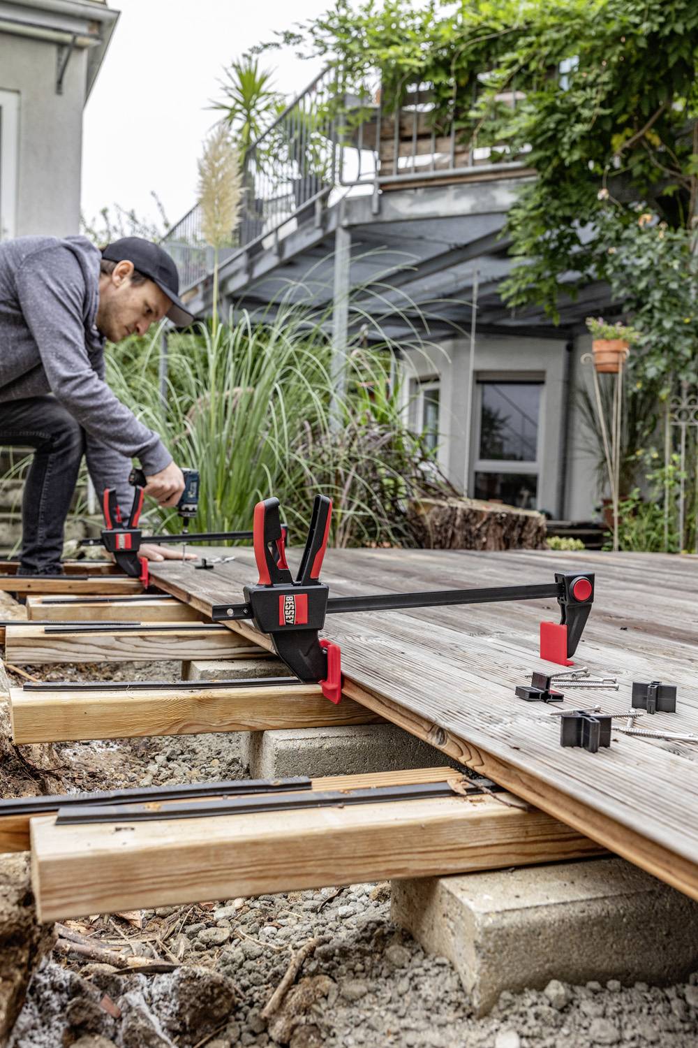 Eine Person montiert Holzterrassenplatten im Garten mit Klemmen und Werkzeugen, um die Konstruktion stabil und eben zu sichern.