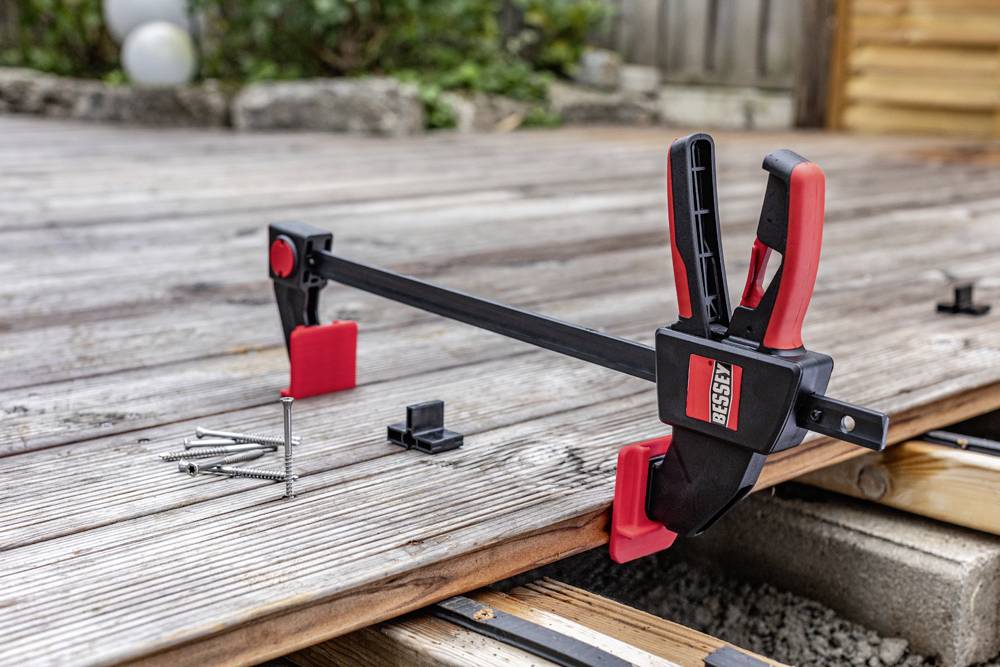 Auf einer Holzterrasse liegt eine Spannzwinge neben Schrauben und Clips. Die Terrasse wird offenbar renoviert oder gebaut.