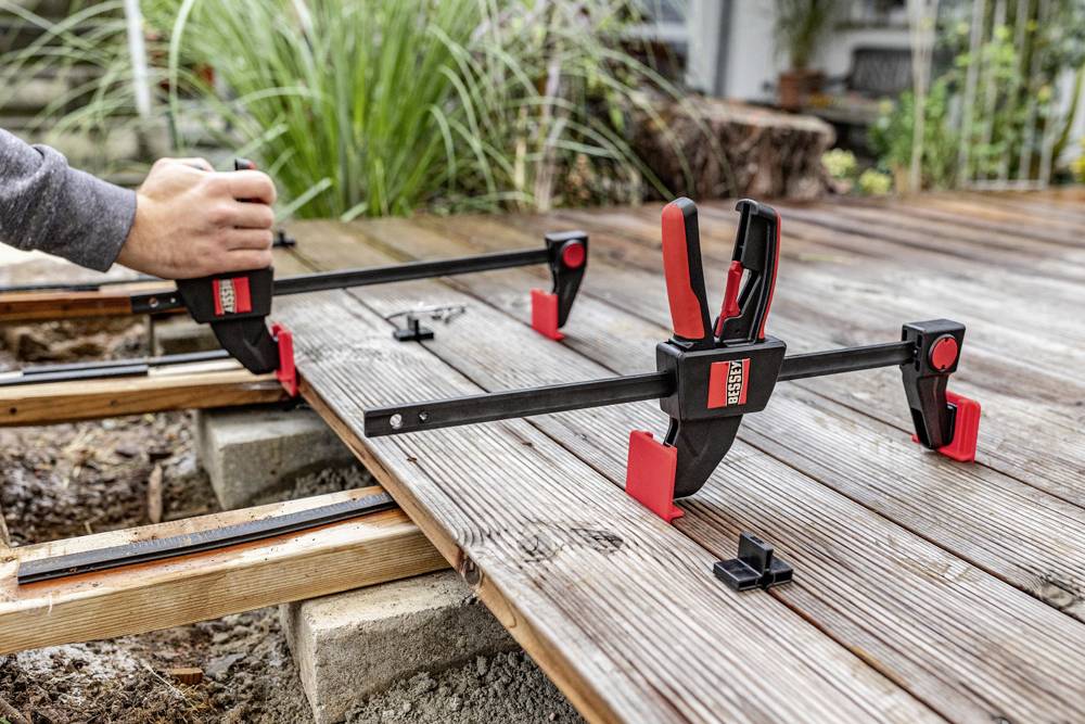 Eine Person verwendet Schraubzwingen, um Holzbretter auf einer Terrasse zu befestigen. Umgebung zeigt grünes Gras im Hintergrund.